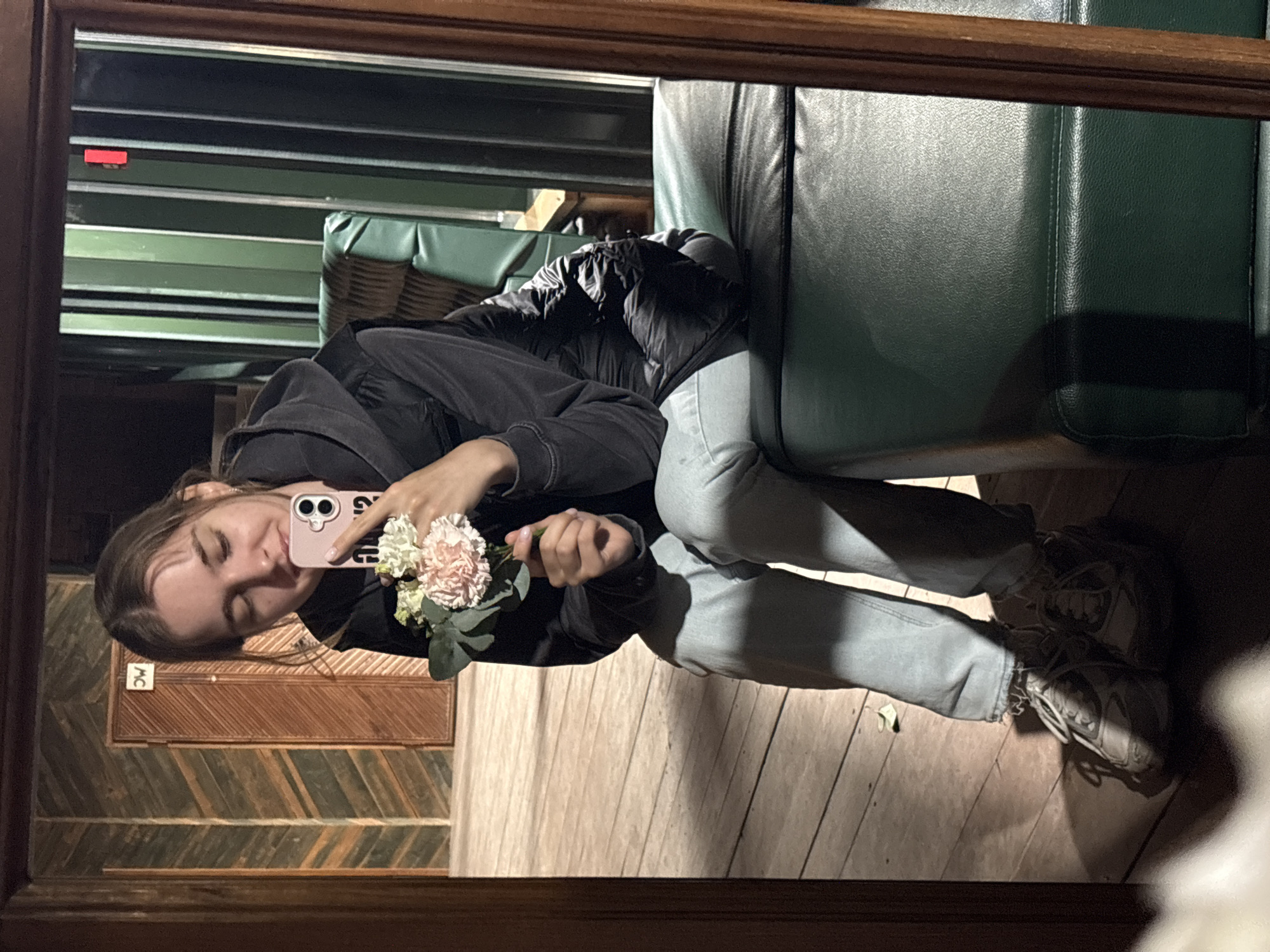 An indoor mirror selfie taken in Kanabekovo, Russia, depicts a young Caucasian female seated on the edge of a dark green cushioned surface. She wears a dark hooded sweatshirt, a dark puffy vest, light blue jeans, and light-colored sneakers. In her right hand, she holds a light pink smartphone with a dual-lens camera, pointed towards herself, indicating a selfie capture. Her eyes are closed, and she exhibits a subtle smile. Her left hand holds a small bouquet of flowers, including pink and white carnations, along with green foliage.
The background, reflected in the mirror, features a wooden wall with a chevron or herringbone pattern on the left, leading to a wooden door with vertical planks. Above the door, an inverted white rectangular sign with dark text is visible. To the right of the subject, a wall with vertical dark green panels is observed, with a red rectangular object affixed near the top of one panel. A dark green cushioned chair with a wicker-style backrest is partially visible behind the subject. The floor is constructed of light brown wooden planks, casting distinct shadows from the subject and objects. A small, dry leaf is visible on the floor in one area. The mirror frame is made of dark wood.