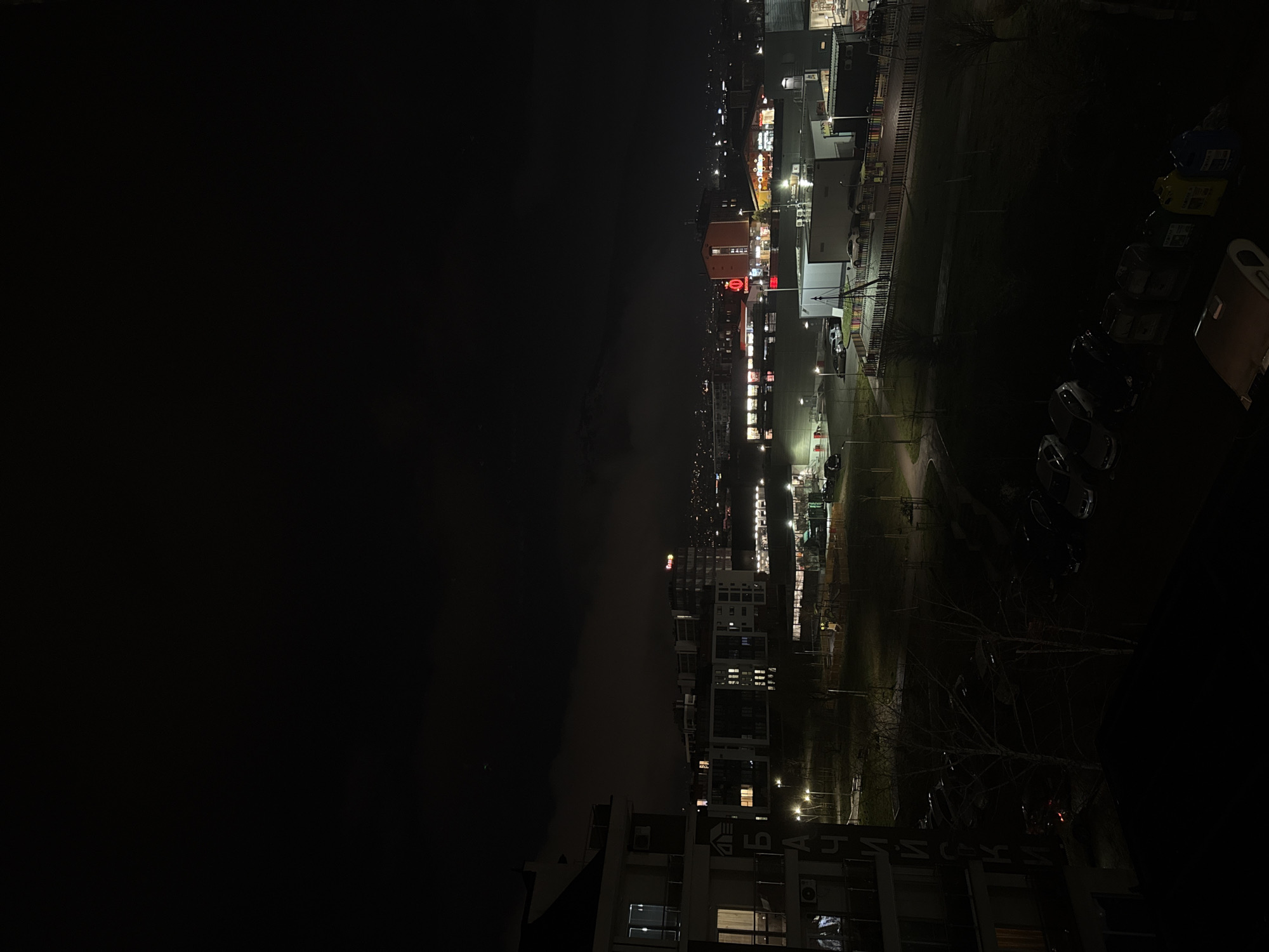 The image depicts a nighttime view of Sofia, Bulgaria, from an elevated position, possibly a high-rise apartment. The right side of the frame shows a brightly lit urban landscape, including commercial buildings, illuminated signs, and streetlights. A row of parked cars is visible. The left side of the image shows a darker area, possibly obscured by weather, with faint outlines of distant buildings and city lights. The sky is overcast. At the bottom, there's a building with what appears to be Cyrillic text.
