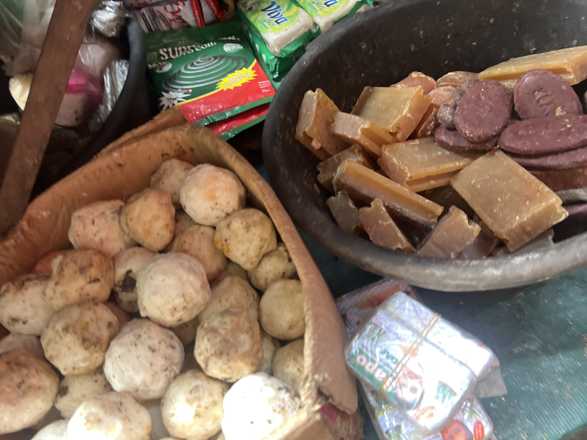 A high-angle, slightly blurry, close-up shot captures a market stall in Nkpor, Nigeria. The scene is filled with a variety of goods, primarily food items and toiletries. In the foreground and to the right, a dark, rough-textured bowl overflows with rectangular blocks of amber-colored soap or shea butter and several round, dark red objects that appear to be cosmetic soaps or medicinal products. Above and to the left of the bowl, a woven basket contains a pile of round, off-white, earth-toned tubers, possibly yams or cassava, some of which have visible dirt specks. To the left of the basket, several wrapped packets of soap, tied with rubber bands, are visible. In the background, green and red packaging for mosquito repellent coils and other toiletries can be seen. The overall impression is of a bustling, informal market environment.