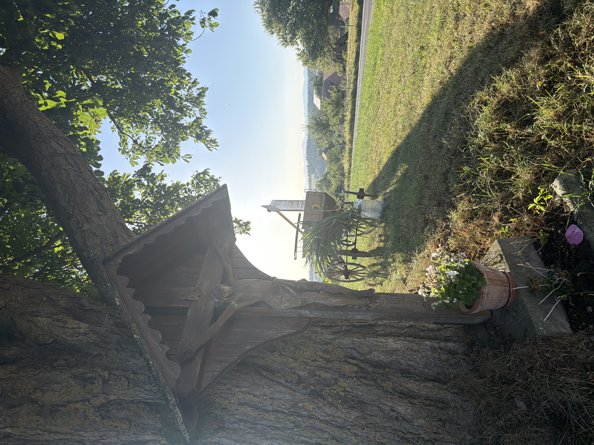 The image depicts a roadside shrine with a wooden crucifix attached to a large tree. The crucifix features a depiction of Jesus Christ. In the background, a vintage wagon with large wheels is visible, along with some greenery and a distant landscape of hills and houses under a clear blue sky. A potted plant with white flowers sits at the base of the tree. The scene appears to be in a rural setting, possibly on the outskirts of Ufhusen, Switzerland, given the presence of traditional rural elements and the natural landscape.
