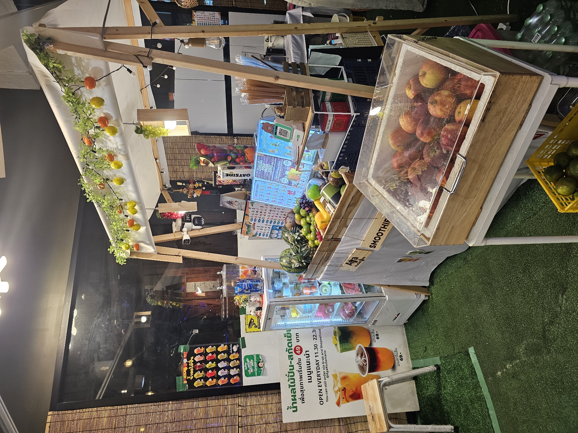 The image shows an outdoor juice and smoothie stand in Bangkok, Thailand. The stand is set up with a wooden frame and a white canopy, decorated with artificial fruit and string lights. In front of the stand is a cooler filled with apples and a basket of oranges. There is a menu board in Thai advertising smoothies and juices. A small refrigerator unit is visible behind the counter, stocked with drinks and fruits. The ground is covered with artificial grass. The overall setting suggests a casual, vibrant atmosphere for a food vendor.