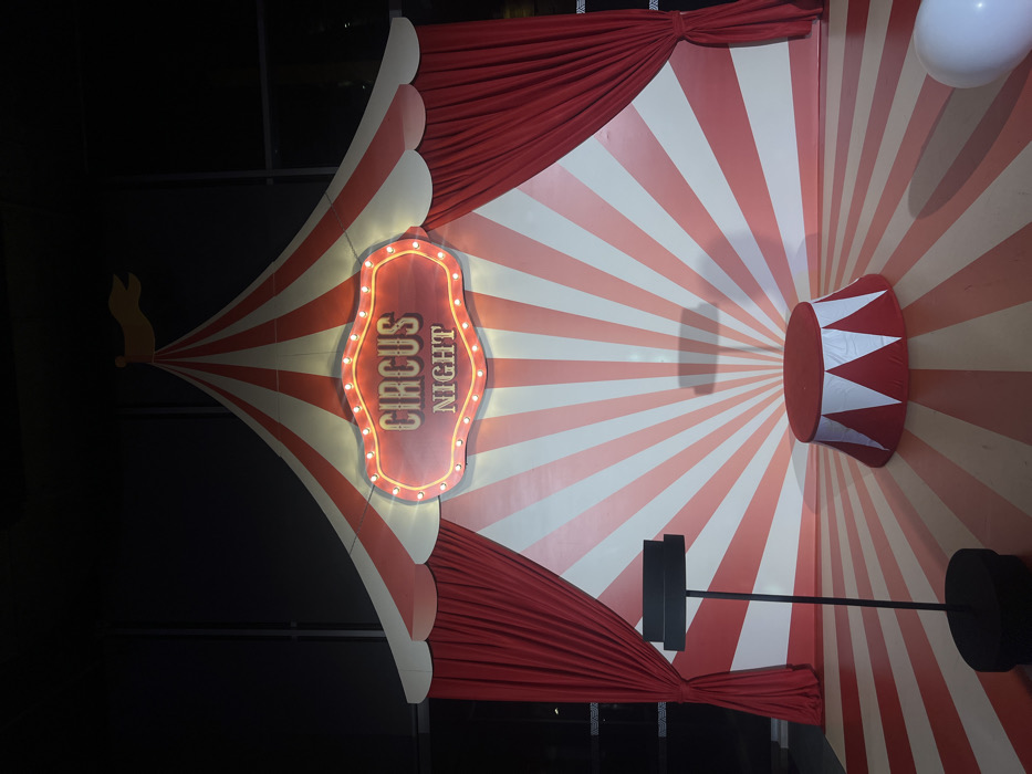 The image displays an unoccupied indoor event setup in Budva, Montenegro, designed with a circus theme. The central feature is a red and white striped backdrop shaped like the upper portion of a big top tent, with radial stripes converging at the top. A small, light yellow pennant flag is visible at the apex of the tent structure. Below this, an illuminated sign with an ornate border and multiple bulbs reads "CIRCUS NIGHT" in two lines against a red background. Red drapes are pulled back and tied on both the left and right sides, framing the striped backdrop. The floor extends the red and white radial stripe pattern from the backdrop towards the foreground. A cylindrical platform, red on top with red and white triangular stripes on its sides, rests on the striped floor to the right of the center. To the left, a tall, slender black stand with a flat, rectangular top is positioned on the floor, casting a shadow on the striped wall. A white balloon is partially visible in the lower right corner of the frame. No individuals are present, and no actions or interactions are depicted.