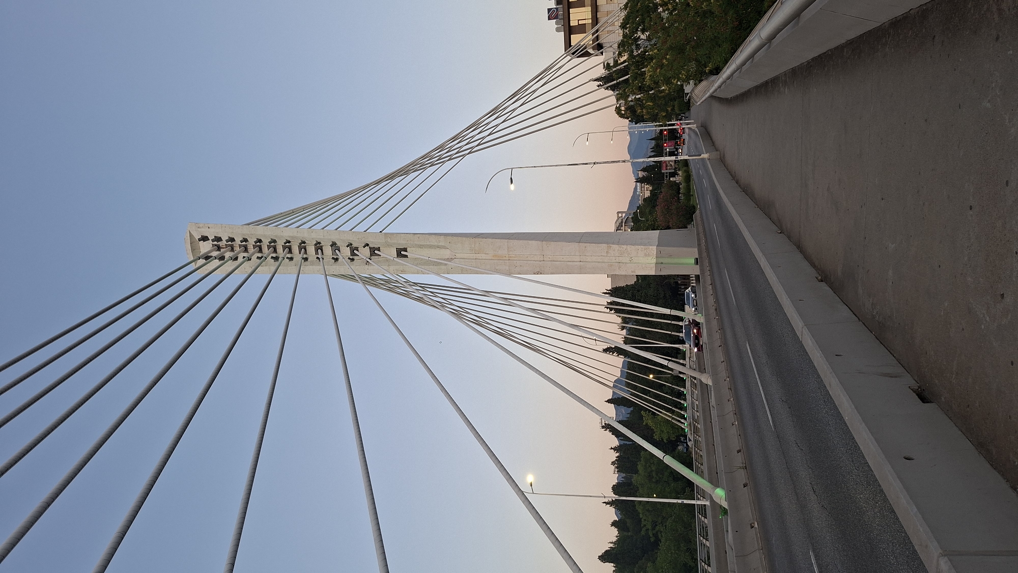 This is a full shot of the Millennium Bridge in Podgorica, Montenegro, captured from the pedestrian walkway on the right side of the bridge. The image is taken at dusk or dawn, as indicated by the soft, warm light and the clear, pale blue sky. The bridge's iconic white pylon stands prominently in the center, with numerous cables fanning out to support the road deck. The road itself is visible, with two cars driving away from the camera. A sidewalk runs alongside the road, with concrete barriers separating it from the traffic and the bridge structure. Green lights illuminate parts of the bridge's base, and trees and buildings can be seen in the background, extending to distant mountains.