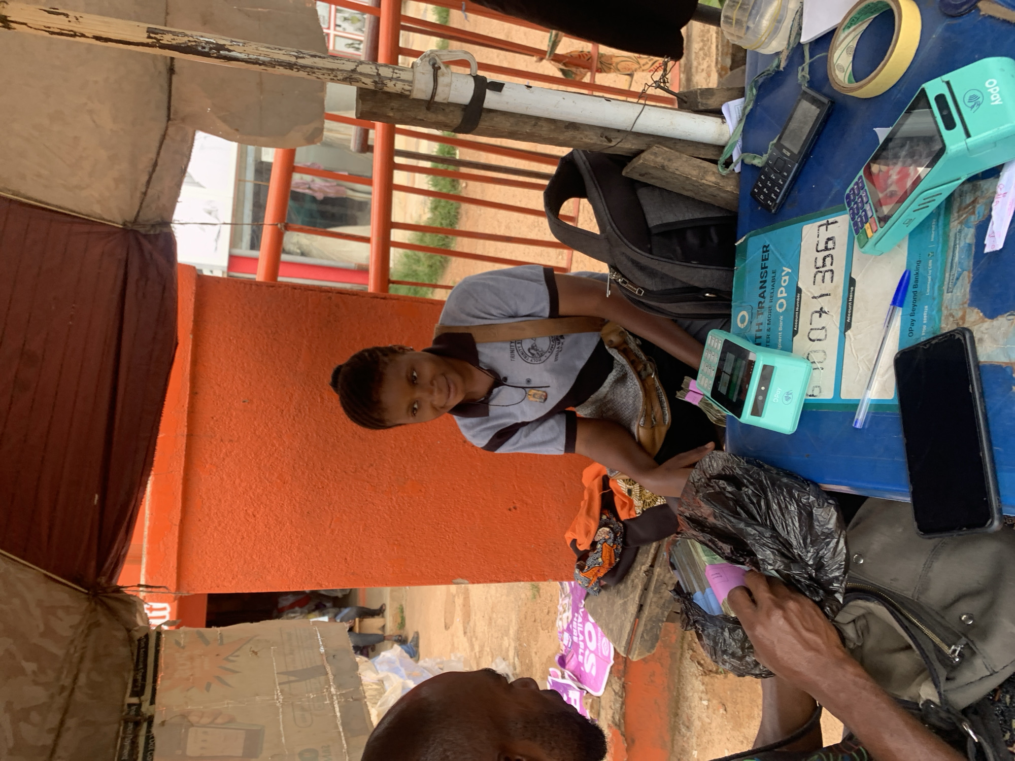 The image, taken in Jos, Nigeria, captures an outdoor financial transaction at a makeshift stall during daytime. A smiling young woman, wearing a grey polo shirt with "TRINITY" visible on it, stands behind a blue plastic table. She is interacting with a male customer, whose head and arm are visible in the foreground, holding wads of cash within a black plastic bag. This suggests a payment, deposit, or withdrawal transaction.
On the blue table, several items essential for financial services are laid out. Two distinctive light blue/green OPay Point-of-Sale (POS) terminals are prominently displayed. Alongside them are an older model black mobile phone, a roll of yellow adhesive tape, and a blue pen. A sign detailing OPay transfer services is visible, with text including "OPay," "TH TRANSFER TER & MORE RELIABLE," and sections for "Account Number" and "Account Name." The account number "6700713567" is clearly legible. Additional OPay branding, "OPay Beyond Banking..." and "Licensed by CBN," is also present.
The setting indicates an informal street or market environment, with an orange textured wall in the background and a simple structure of wooden and metal poles supporting a canopy or stall. The bright, clear lighting suggests it is a sunny day. The overall scene depicts a common financial service provided by a POS agent in a bustling urban context.