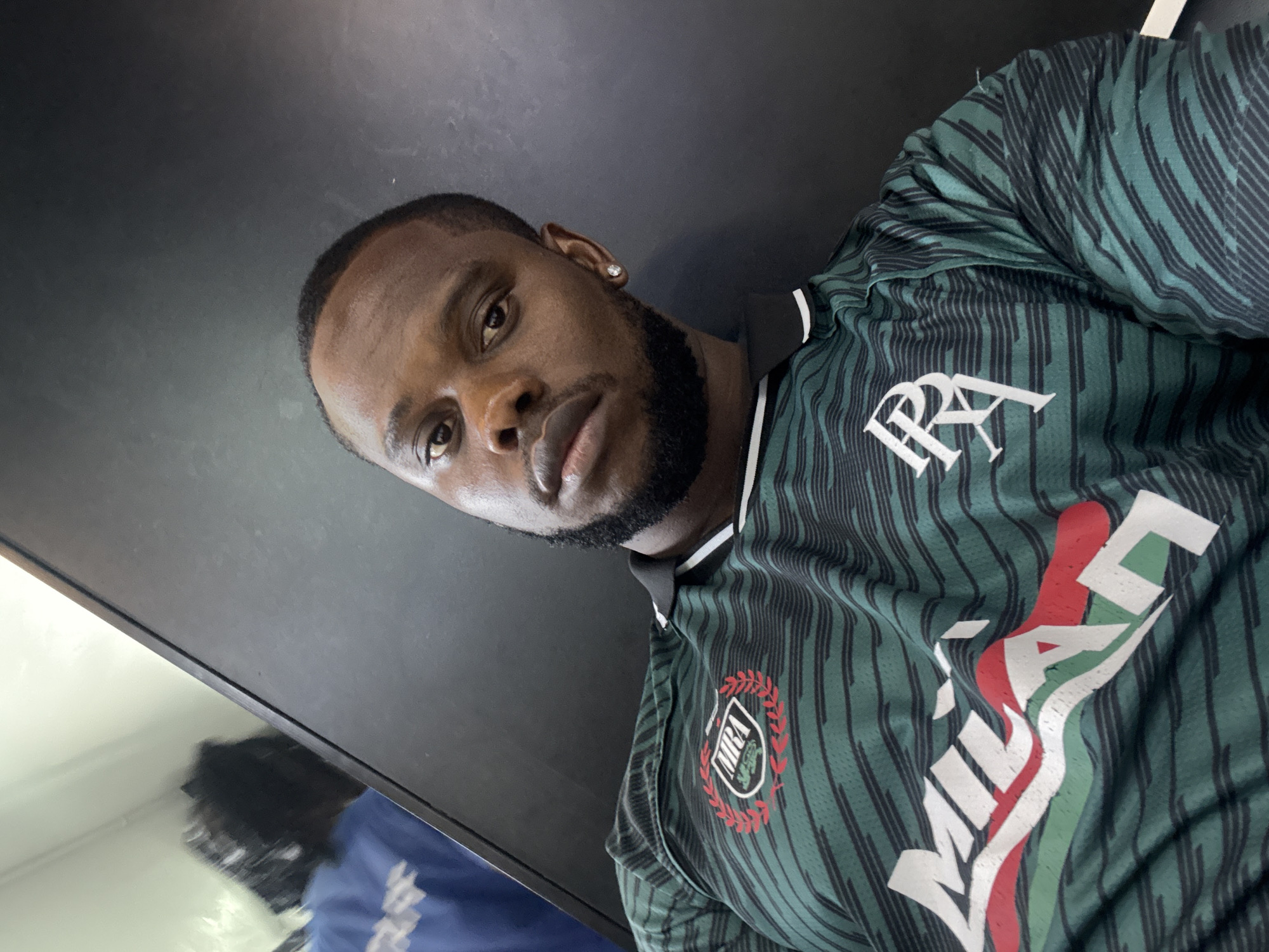 A man with a beard and short, dark hair is wearing a green and black striped Milan jersey, with "MRA" logos on the jersey and "RA" above the "Milan" wording. He is wearing a small earring in his left ear. The man appears to be standing inside a mirrored elevator or a small mirrored room with dark walls. The location is Lugbe, Nigeria.