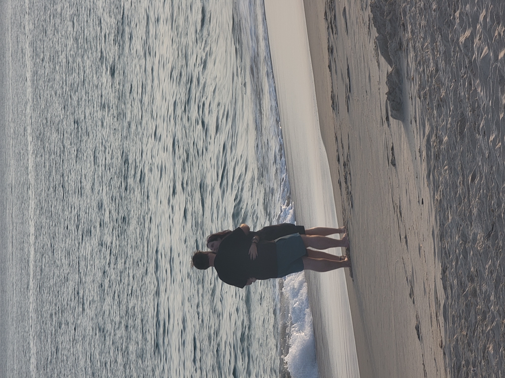 A man and a woman are embracing on a sandy beach in Quarteira, Portugal. The man is wearing a dark t-shirt and blue shorts, while the woman is wearing a dark dress. They are standing near the water's edge, with waves gently lapping at the shore. The ocean stretches out behind them, a calm expanse of rippling water under a soft, diffused light. The sand is textured with small footprints and ripples, suggesting a tranquil, natural environment.