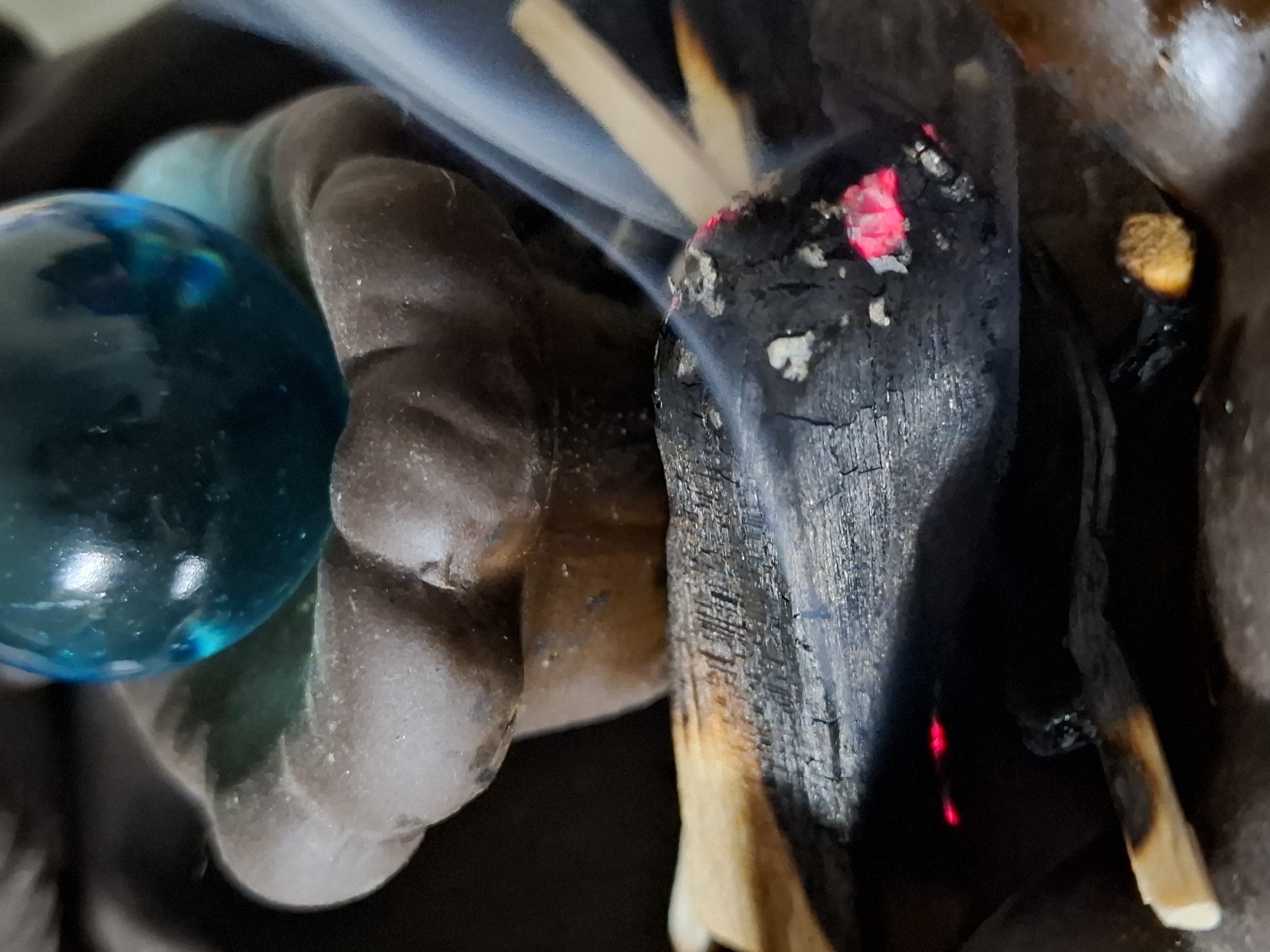 The image shows a close-up of a hand holding a burning piece of wood or incense stick. The wood is charred and glowing red at the tip, with smoke rising from it. A blue glass ball rests on the hand. The background is blurred. It's impossible to determine a location based on this image.