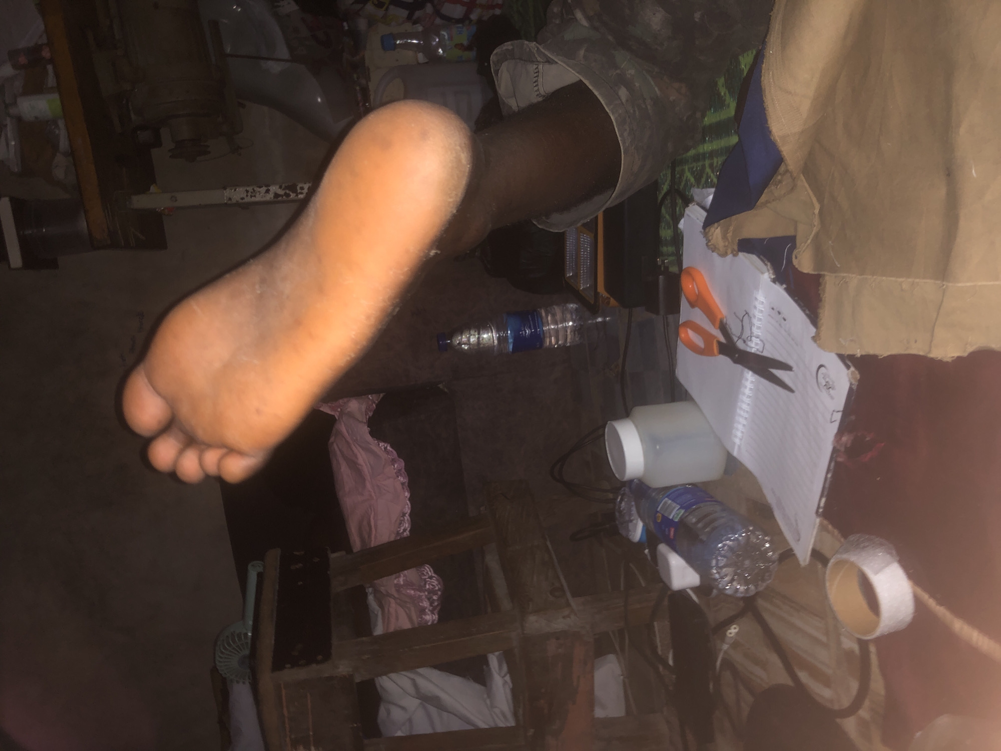 The image depicts a dimly lit indoor scene, likely a residential area in Ado, Nigeria. The focal point is a person's bare foot, prominently displayed in the foreground, with visible skin texture and color. Surrounding the foot are various objects scattered across a surface, possibly a bed or floor. These include a plastic water bottle, a white jar with a lid, a notebook with a pen, and a pair of orange-handled scissors. Further in the background, a wooden stool with what appears to be fabric draped over it is visible, as is a sewing machine and other assorted items, suggesting a living or workspace. The overall impression is one of a cluttered yet functional environment.
