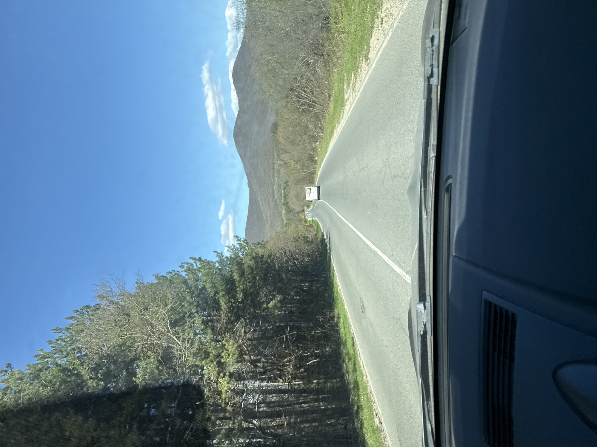 The view is from inside a vehicle, looking out the windshield onto a paved road, likely in Pryaporets, Bulgaria. The road extends straight ahead with a white dividing line. A white box-shaped vehicle is visible some distance down the road. The road is bordered on the left by trees and vegetation and on the right by a mountain in the distance. The sky is blue with some scattered clouds. The interior of the vehicle is visible on the right side of the frame.