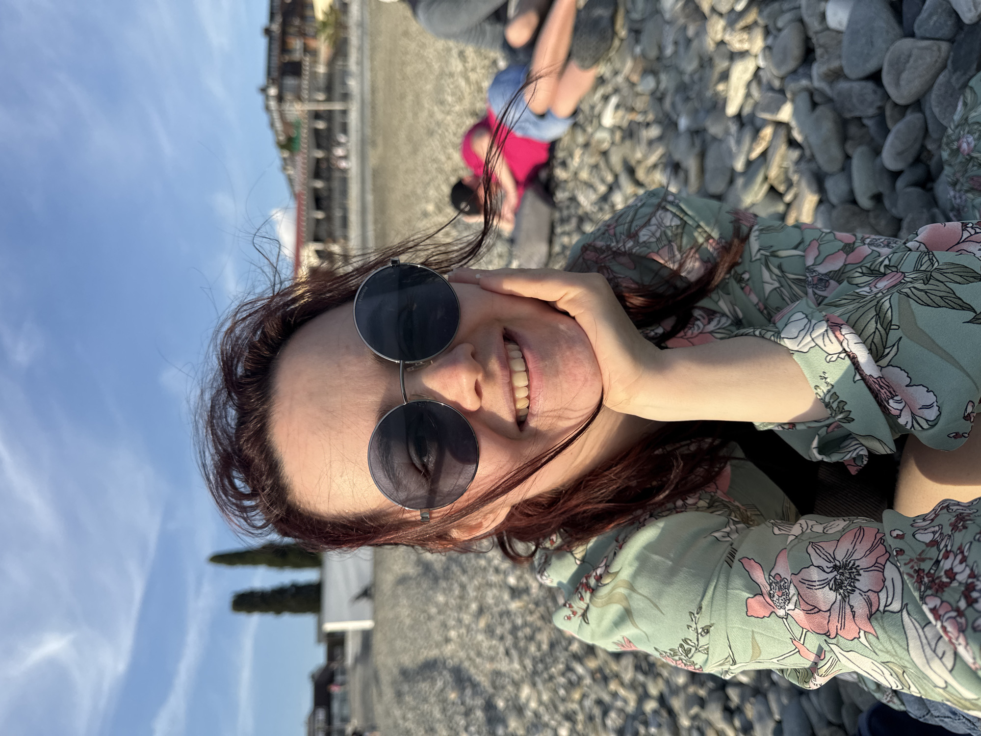 A woman with red hair is shown from the chest up in the foreground, wearing sunglasses and a light green floral blouse. Her right hand is touching her face. In the background are other individuals, pebbles, buildings, trees, and a blue sky. The photo was taken in Sochi, Russia.