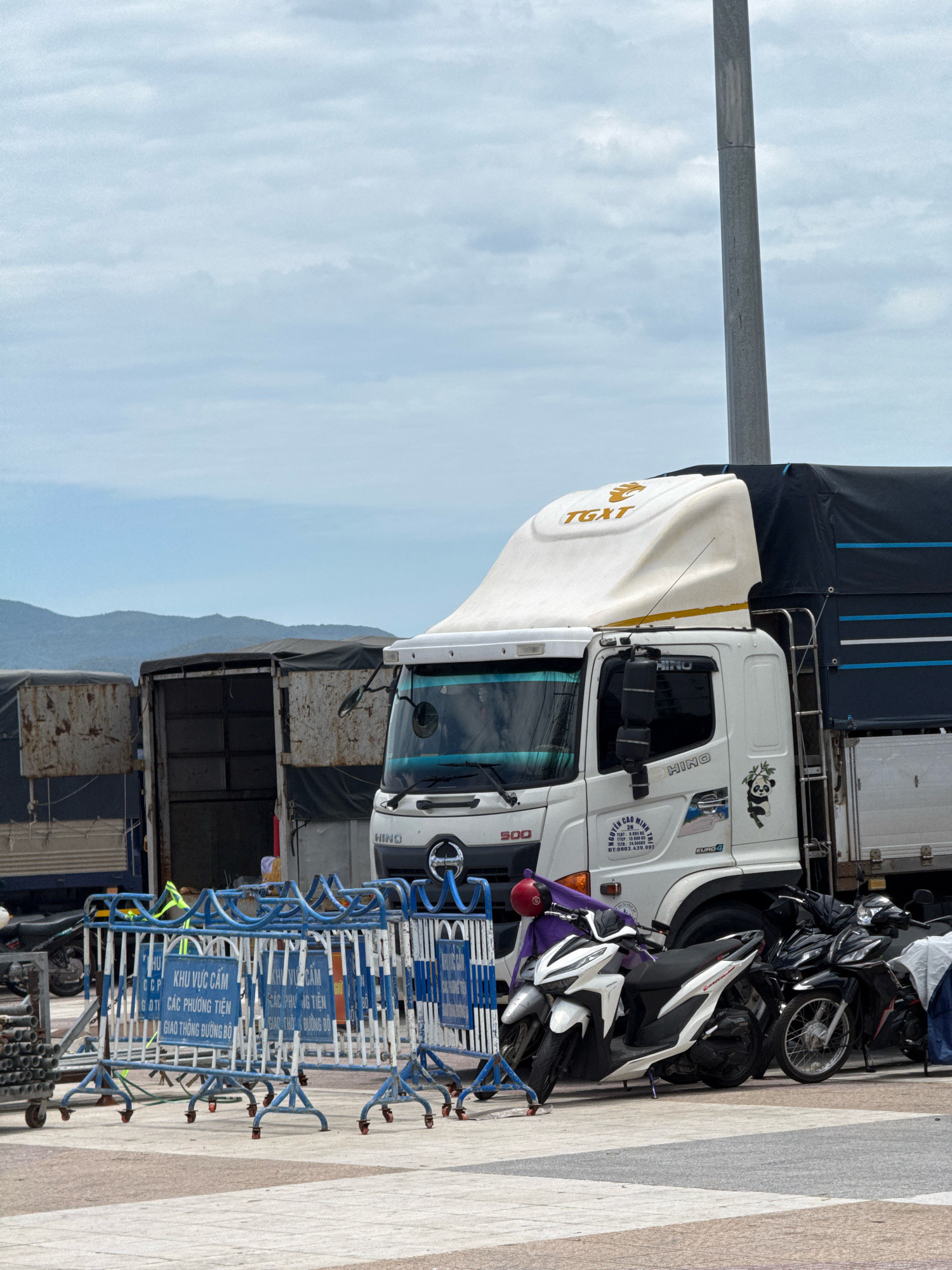 The image shows a white Hino truck parked next to other trucks and several motorcycles. The truck has a black tarp covering its cargo bed, and a white aerodynamic fairing on its roof with the text "TGXT" in gold. A graphic of a panda is visible on the side of the truck's cab. In the foreground, blue and white barricades are set up, with signs in Vietnamese that appear to indicate a restricted area and traffic restrictions. Two motorcycles, one white and one black, are parked behind the barricades. The scene appears to be an outdoor area with a paved surface, and a cloudy sky with distant hills in the background. The presence of Hino trucks, the Vietnamese text on the signs and truck, and the overall aesthetic suggest the location is in Vietnam. Given the context of a port or transportation hub, and the commonality of such scenes in coastal cities of Vietnam, this could be the port area of Nha Trang.
