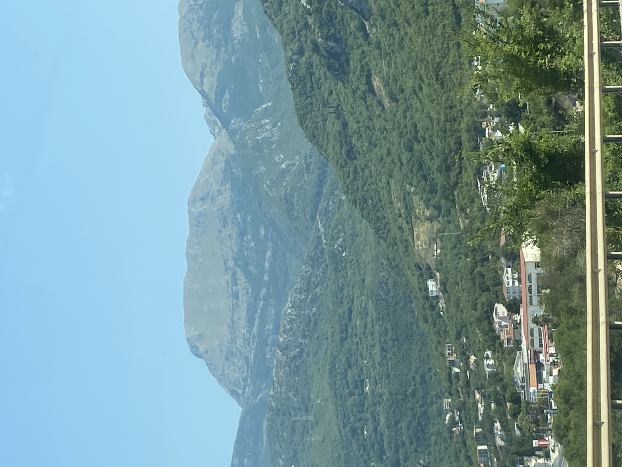 The image depicts a mountainous landscape in Prijevor, Montenegro. In the foreground, a metal railing is visible, suggesting the viewer is on a bridge or elevated viewpoint. Beyond the railing, a densely forested hillside slopes upwards, dotted with scattered houses and buildings. Further in the distance, larger, more rugged mountains dominate the horizon under a clear blue sky. The scene is characterized by lush greenery, residential structures built into the hillside, and imposing natural formations. There are no people or animals visible in the image.