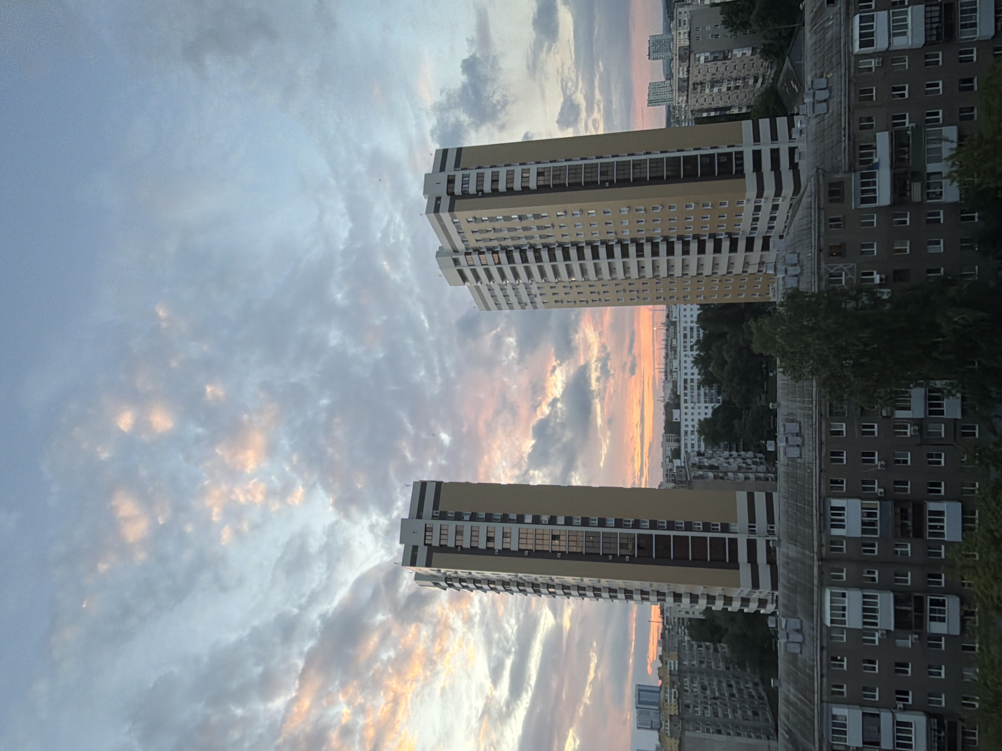 This is an eye-level, exterior shot of a city skyline at dusk. The sky is filled with dramatic clouds, tinged with pink and orange from the setting sun. In the foreground, a gray, multi-story apartment building with many windows and balconies dominates the lower portion of the frame. Behind it, two tall, modern high-rise buildings stand prominently. The left building is primarily tan with brown and white accents, while the right building is a lighter tan with white and darker tan accents. Numerous other apartment buildings and trees are visible in the mid-ground and background, stretching towards the horizon. The lighting suggests it is late evening.