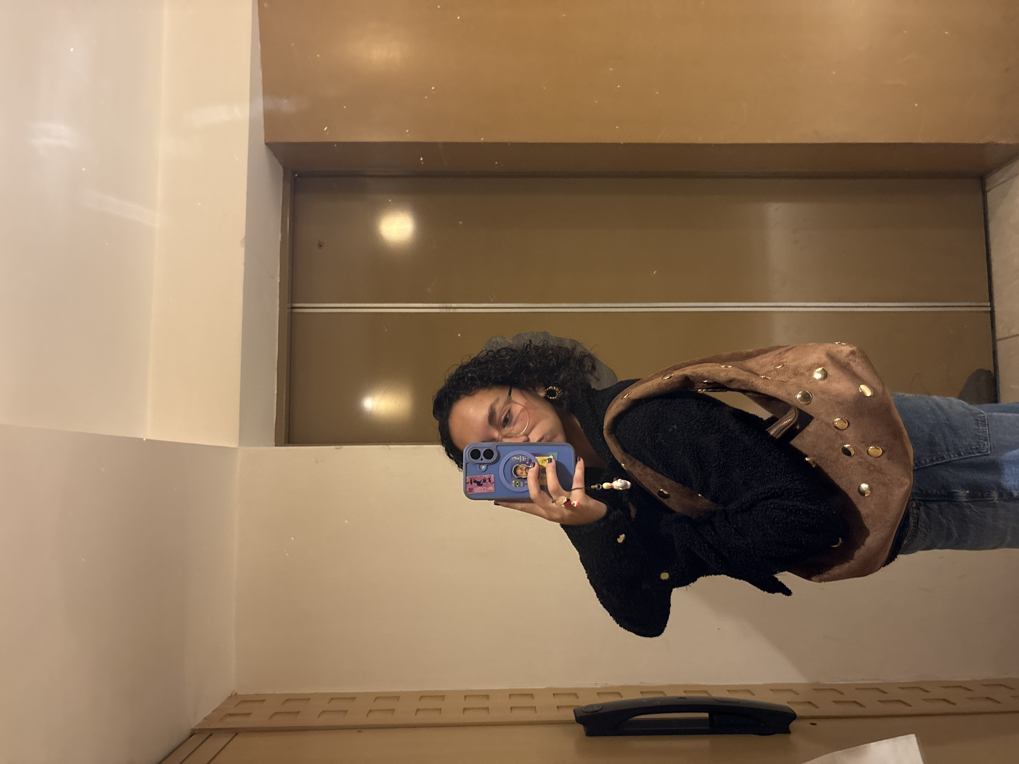 A reflection in a mirror shows a person with curly dark hair, wearing glasses and a black fleece jacket, taking a selfie with a blue phone adorned with stickers. The person is wearing jeans and has a brown, studded purse over their shoulder. The background visible in the mirror is a brown surface with horizontal lines, likely an elevator interior. The surrounding walls appear to be light beige, suggesting an interior setting. The location is likely Mexico City, Mexico. A black object rests near the bottom edge of the frame, possibly a floor mat or cleaning tool.