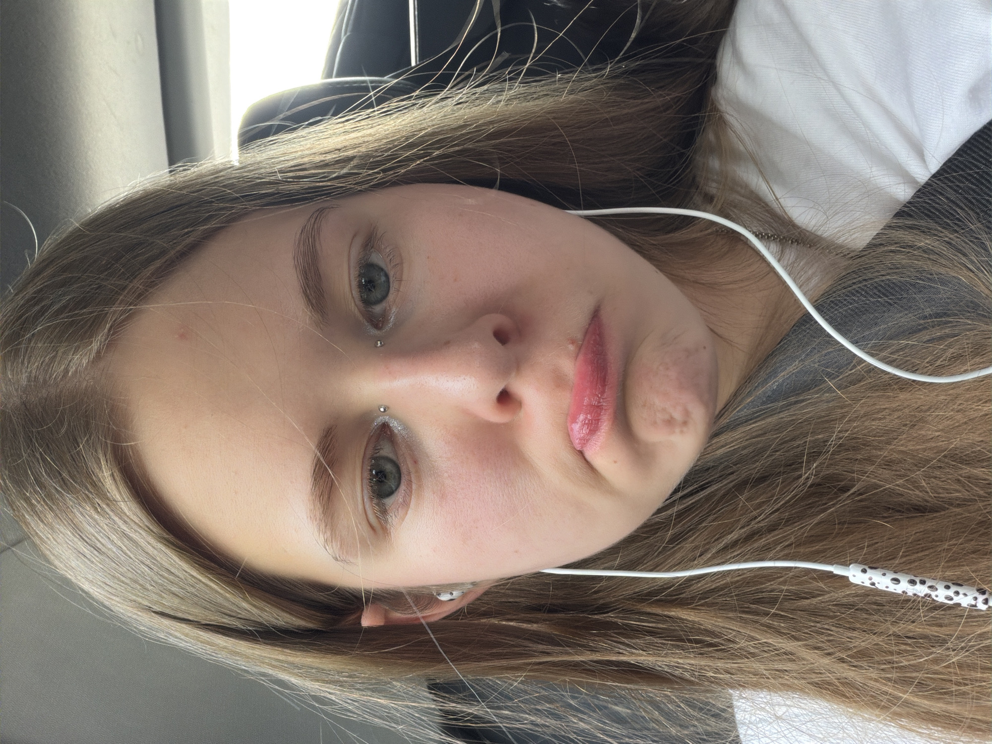 A close-up, waist-up shot captures a young person with long, light brown hair, wearing a white t-shirt and earphones, seated inside a vehicle. The individual's head is tilted slightly, and their facial expression is one of sadness or disappointment, with pursed lips and a downward gaze. A silver eyebrow piercing is visible. The background suggests the interior of a car, with a hint of a headrest and the ceiling. The image does not provide any specific details that would allow for localization in Moscow, Russia.
