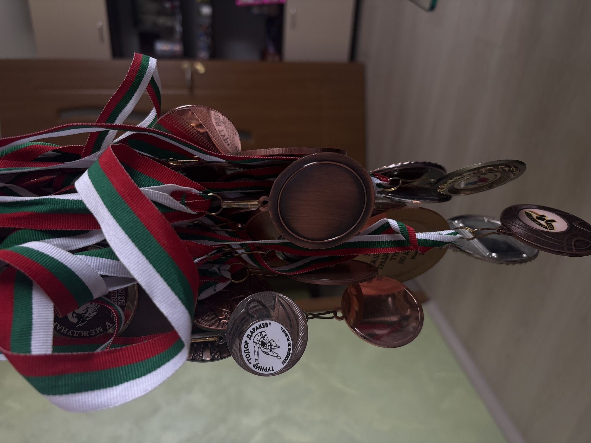 The image shows a collection of medals with ribbons in red, white, and green, the colors of the Bulgarian flag. The medals are displayed on a reflective surface. Close examination reveals inscriptions on some medals, including "ТОДОР ДАРАКЕВ", "ТУРНИР", "ПЛОВДИВ 20.10.2018 г.", "V МЕЖДУНАР", and "2017". The image suggests the location is related to sports achievements, possibly in Plovdiv, Bulgaria. The presence of the Bulgarian flag colors and the city of Plovdiv point to Bulgaria. However, without additional information, determining if the location is in Sofia is not possible.
