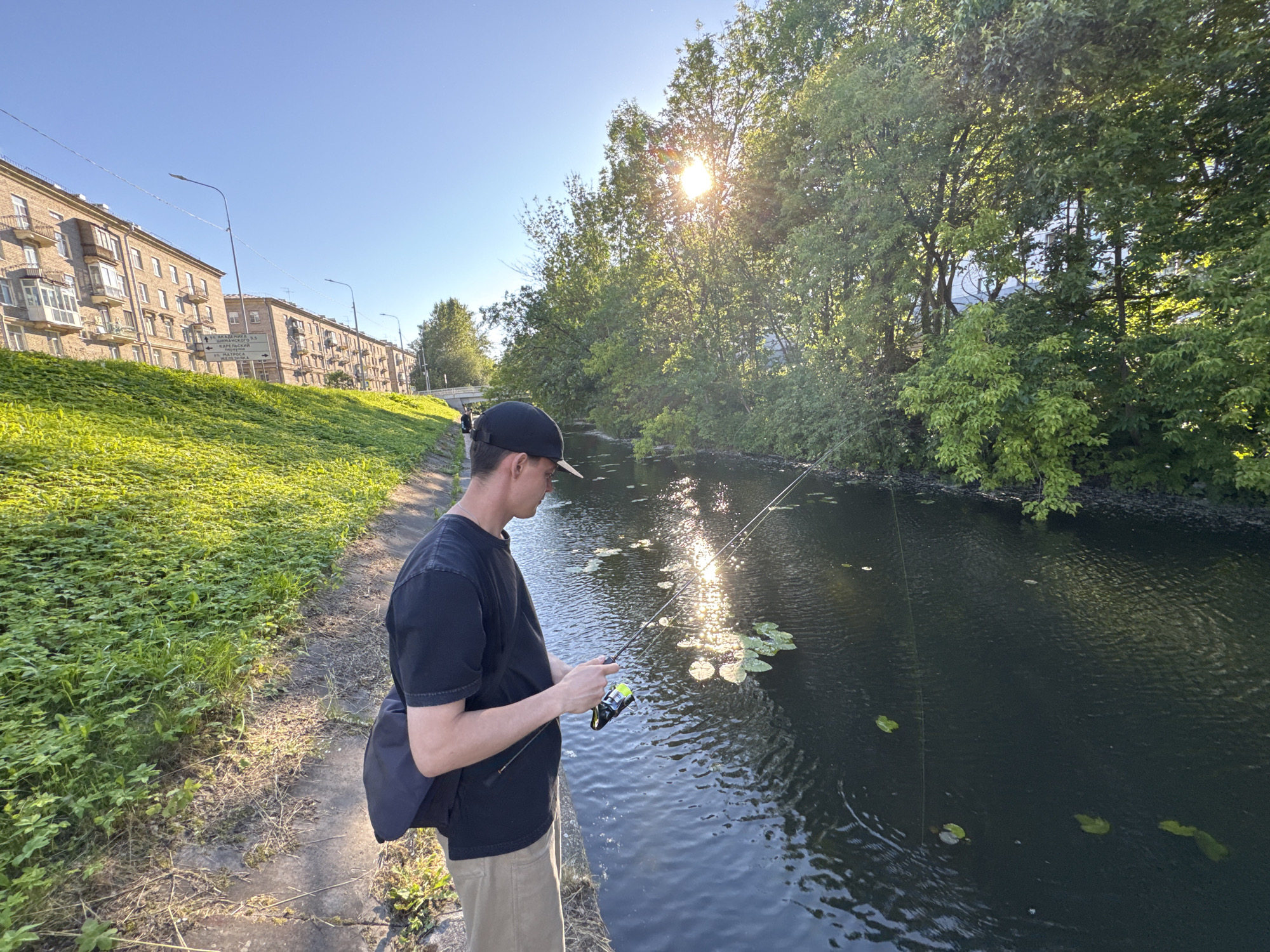 In Saint Petersburg, Russia, on a clear, sunny day, a young man fishes in a canal. He stands on the bank, a black baseball cap on his head, a dark t-shirt, and tan shorts. A dark bag is slung over his shoulder. He holds a fishing rod, actively engaged in the activity. The canal water is dark, reflecting the bright sun and the lush green trees lining its banks. Lily pads dot the water's surface. In the background, apartment buildings with balconies line the opposite bank, and a street lamp can be seen. The sun is high in the sky, indicating it is likely midday or early afternoon. The overall atmosphere is peaceful and serene, with the man absorbed in his hobby amidst the urban greenery. No visible text is present in the image.