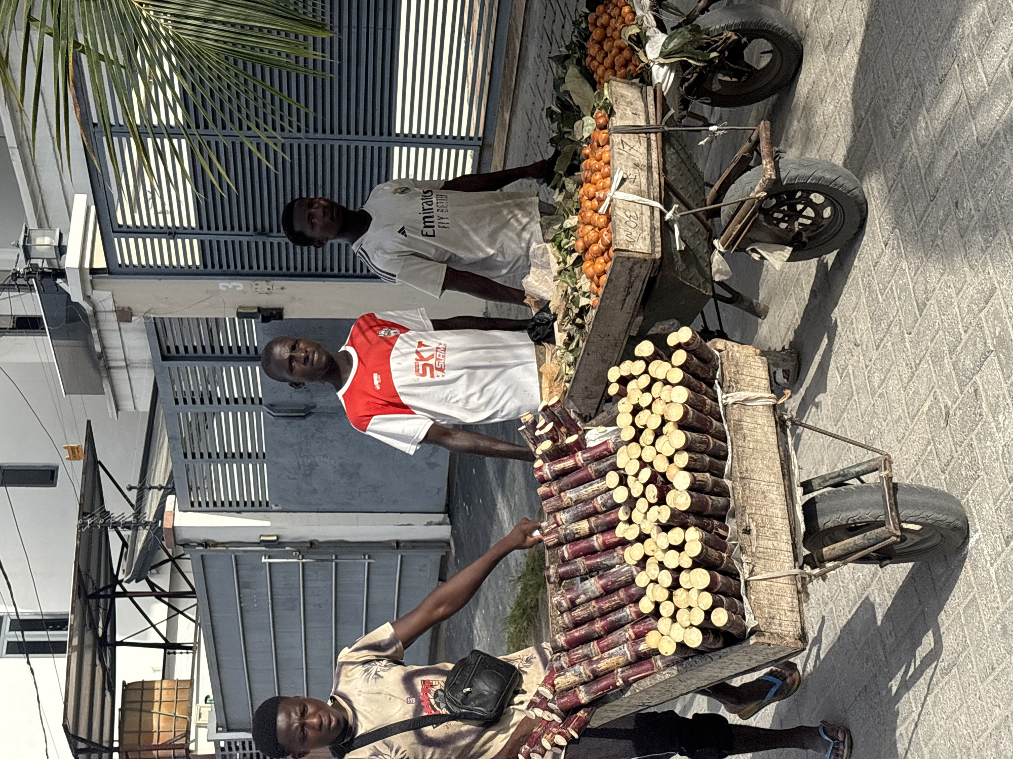 The scene shows three men standing near a wheeled cart in Igboefon, Nigeria. The cart is divided into two sections, one containing stacks of sugarcane and the other holding oranges. One man is pointing at the sugarcane, indicating a transaction or interaction. The two other men stand behind the carts of produce. The backdrop includes a building with closed metal gates. The weather appears sunny.