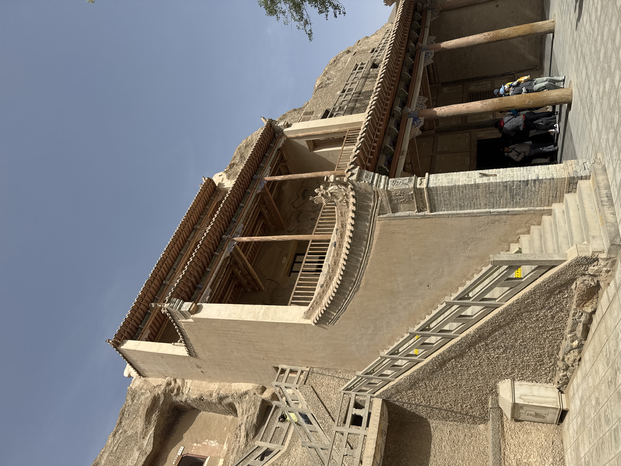The image depicts a building at the Mogao Caves, a UNESCO World Heritage site in Dunhuang City, China. The architectural style is a blend of traditional Chinese and cave-carved elements. The building features a tiled roof, wooden support beams, and a balcony with decorative elements. A set of stairs leads up to the building's entrance. The structure is partially carved into a cliff face. Several people are visible on a covered terrace inside the building. The sky is clear and blue.