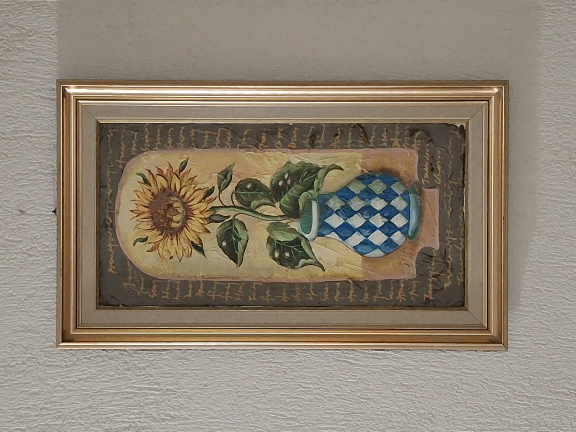 The image shows a framed painting of a sunflower, leaves, and a blue-and-white checkered vase. The painting is rectangular and has a gold frame. The background of the painting is a light beige color with a dark brown border. The painting depicts the sunflower and leaves extending to the left and the vase to the right, the scene appears to be of an interior. There is no information about the location in Krumovgrad, Bulgaria.