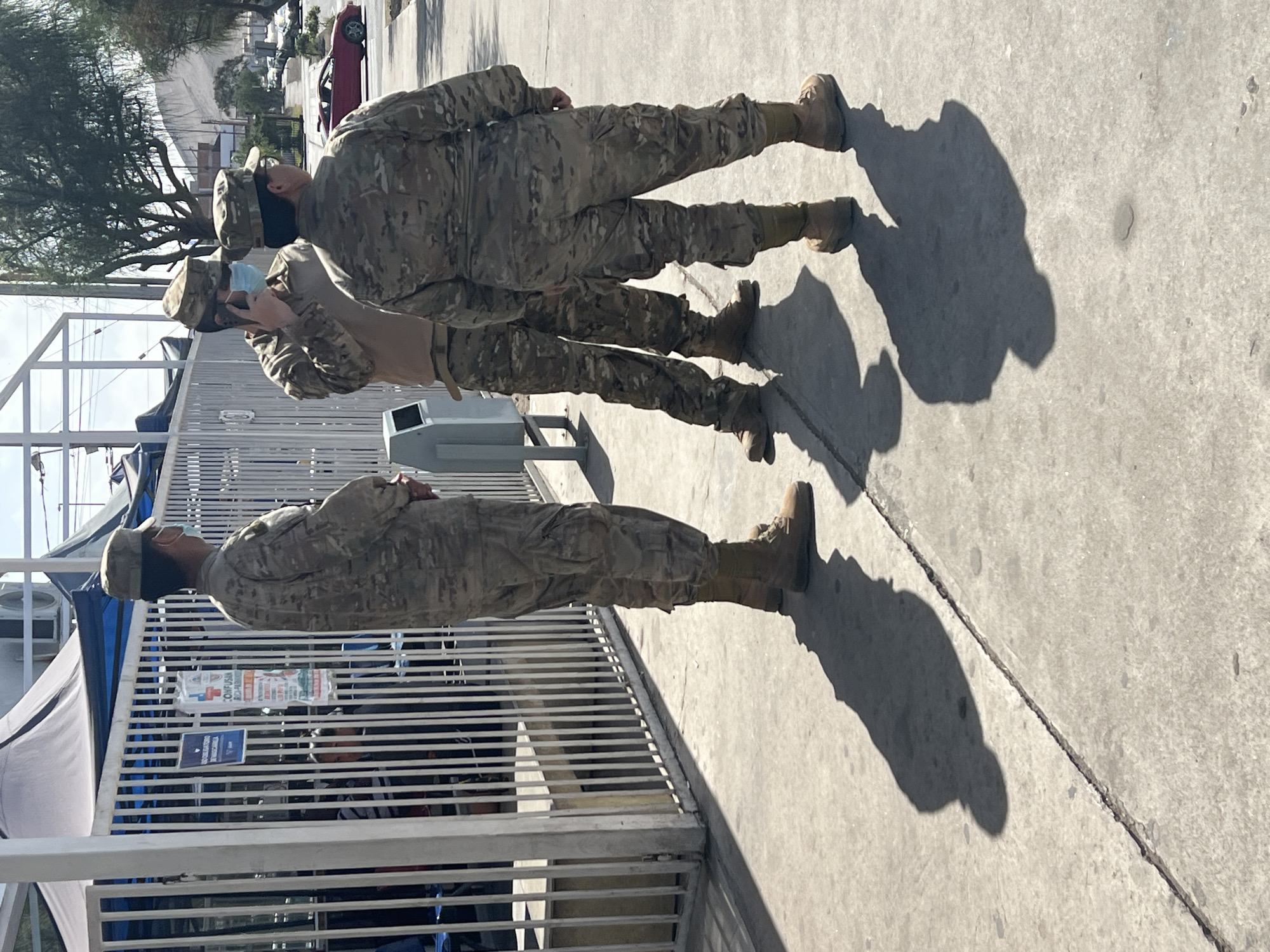 Three individuals in camouflage military uniforms and caps stand on a concrete sidewalk in Arica, Chile. Two individuals are facing away from the camera, and one is in profile, looking to the left. The individual in profile is wearing a light blue face mask and holding a cell phone to their ear. Behind the individuals is a white metal fence, a large tent, and some trees. In the background, a red sports car is visible on a street. The scene appears to be outdoors in daylight.