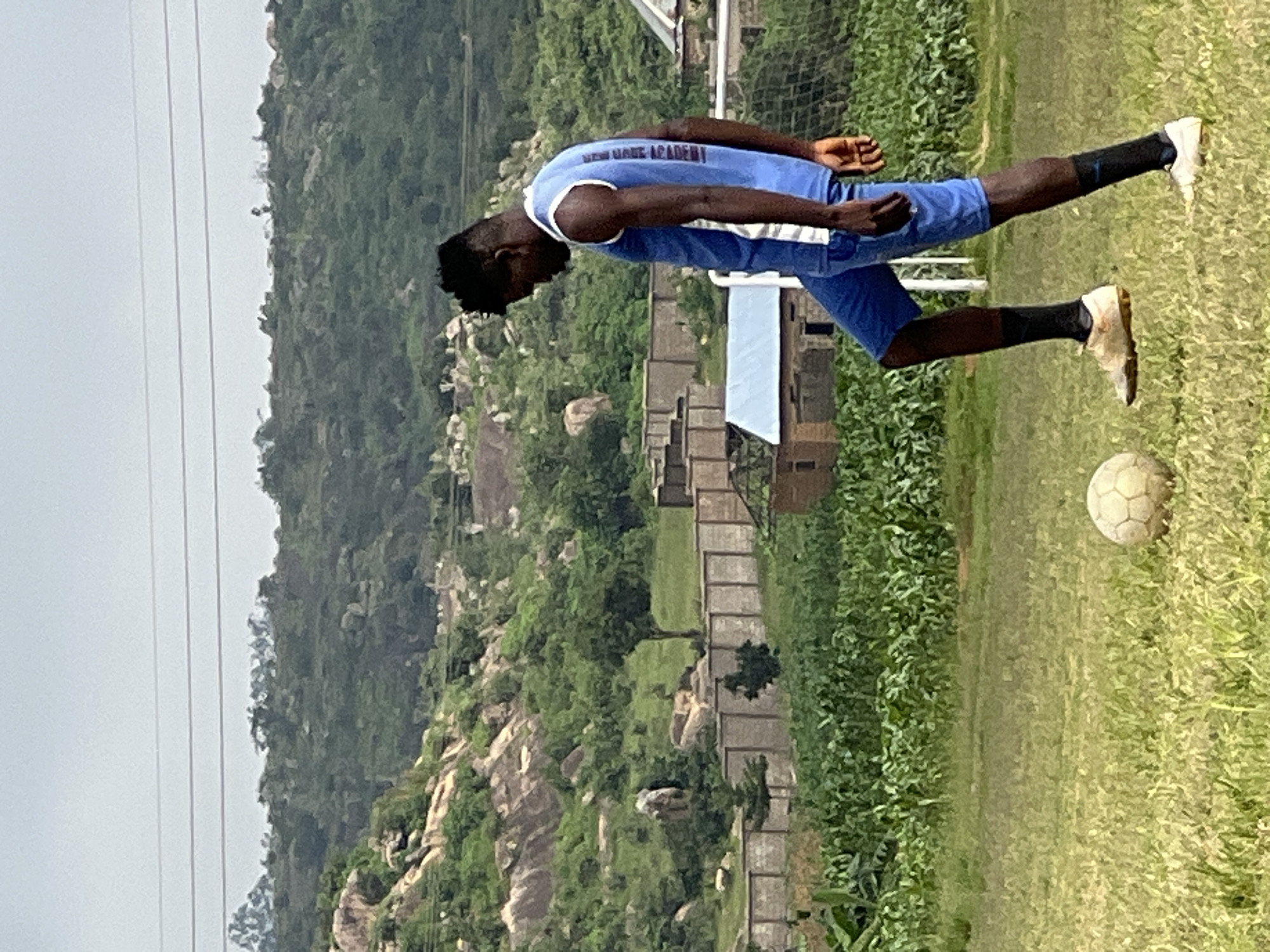 A dark-skinned young man wearing a blue sleeveless jersey with white trim and blue shorts is playing soccer on a grassy field. His jersey has white text on the back. He wears white soccer cleats with black socks. A worn, off-white soccer ball rests on the grass in front of him, positioned beneath his right foot. The background features a lush, green, hilly landscape with scattered rocks and trees. In the mid-ground, a low, light-colored concrete wall runs horizontally across the scene, partially obscuring some buildings and a soccer goal with a white net. A field of green crops, possibly corn, is visible between the wall and the soccer field. The sky is overcast and a pale gray. The overall scene appears to be an outdoor sports or training area on the outskirts of Jos, Nigeria, characterized by its natural hilly terrain and agricultural surroundings.