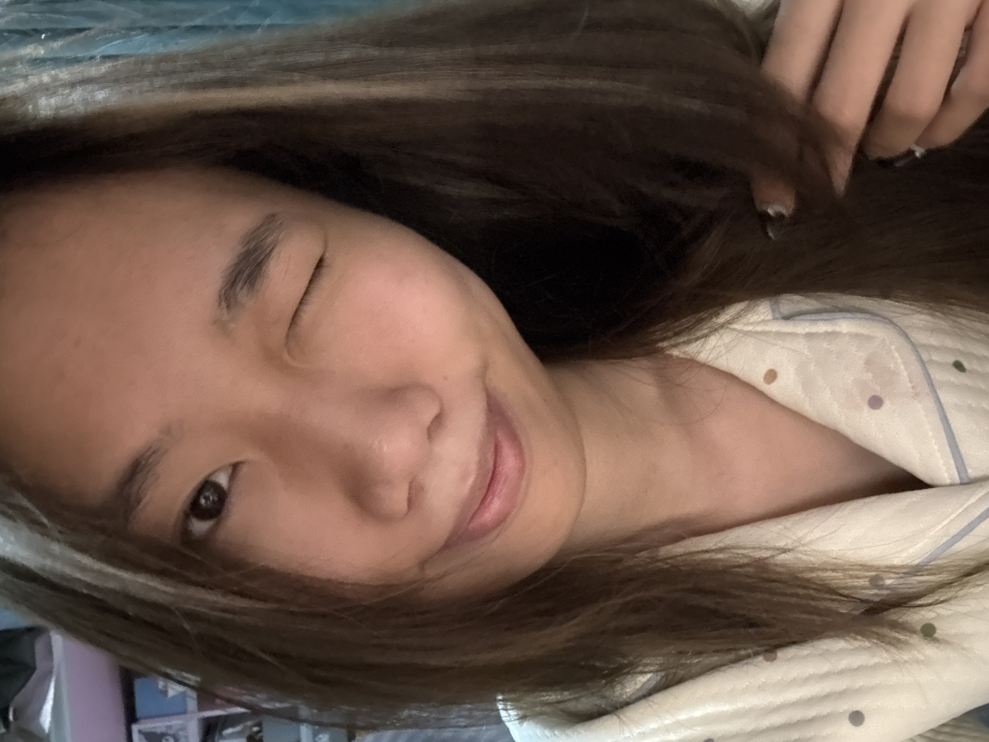 A close-up selfie of a young woman with brown hair. She is winking her left eye and smiling slightly. She is wearing a light-colored pajama top with polka dots and a blue stripe. Her hand is visible in the upper right corner, holding a strand of her hair. A shelf with objects is visible in the blurred background. The specific location within the Nan'an District of China cannot be determined from the image.
