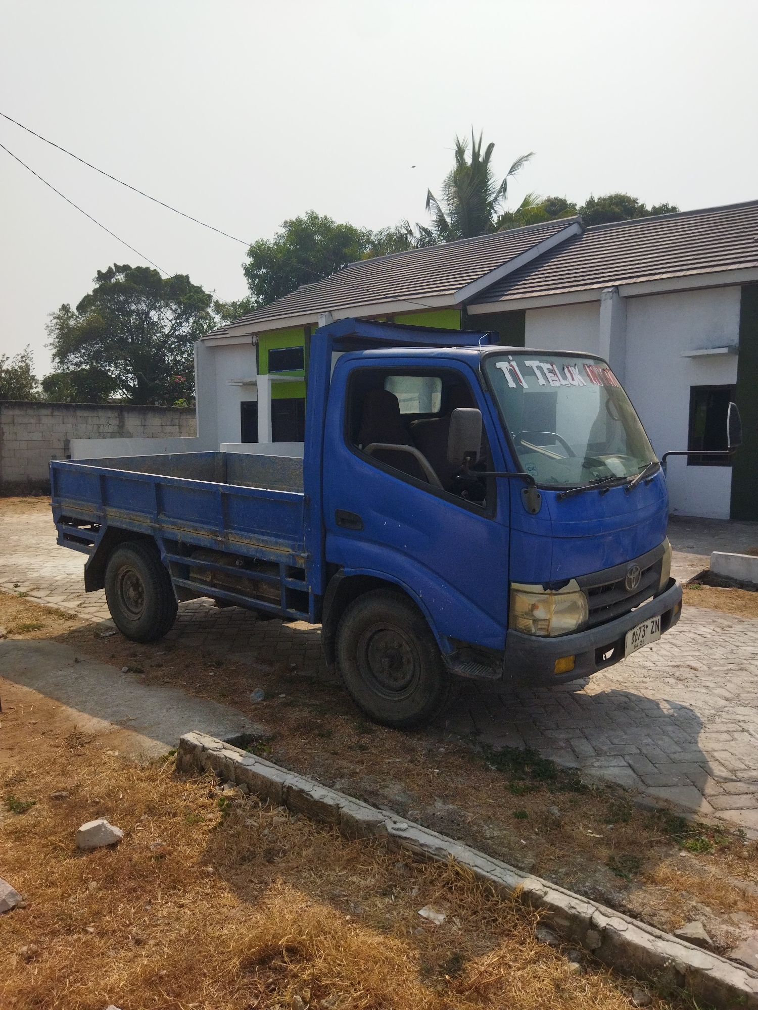 An eye-level, full shot captures a blue, four-wheeled flatbed truck parked on a paved driveway in front of a residential building. The truck, a Toyota Dyna, faces slightly to the right, its bed empty and its driver's side door ajar. The front of the truck displays a license plate reading "B 673 ZN." A white banner with red text, "TI TELON MOCHI," is affixed to the windshield.
Behind the truck, a modern, two-story residential building with a tiled brown roof and white exterior walls is visible. A portion of the building features a bright green accent wall. The yard in front of the building is covered in dry, yellow grass and scattered rocks. A low, concrete border runs along the edge of the driveway. In the background, a dense cluster of trees and a palm tree rise against a clear, pale sky. The overall scene is brightly lit by sunlight. This scene is situated in Suka Sari, Indonesia, a location characterized by its residential areas and tropical climate, suggested by the palm tree and dry vegetation.