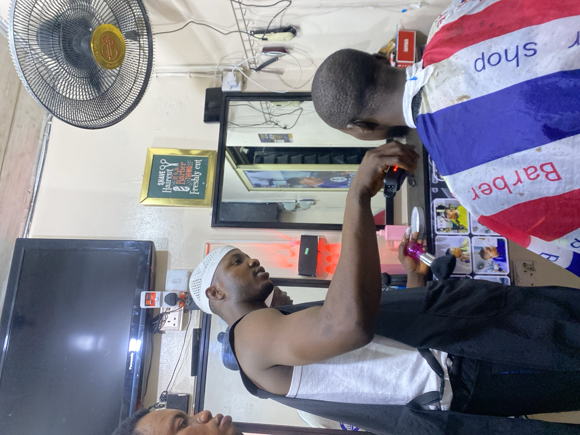In a barbershop in Ado, Nigeria, a barber is giving a haircut to a client. The barber, wearing a white cap, is holding a hair trimmer. The client is draped with a red, white, and blue barber cape. A mirror, a television, an electric fan, and a sign that reads, "Shave, Haircut, It's A Barber Thing, Freshly Cut," are visible in the background. Hair clippers are plugged into the wall outlet above the mirror.