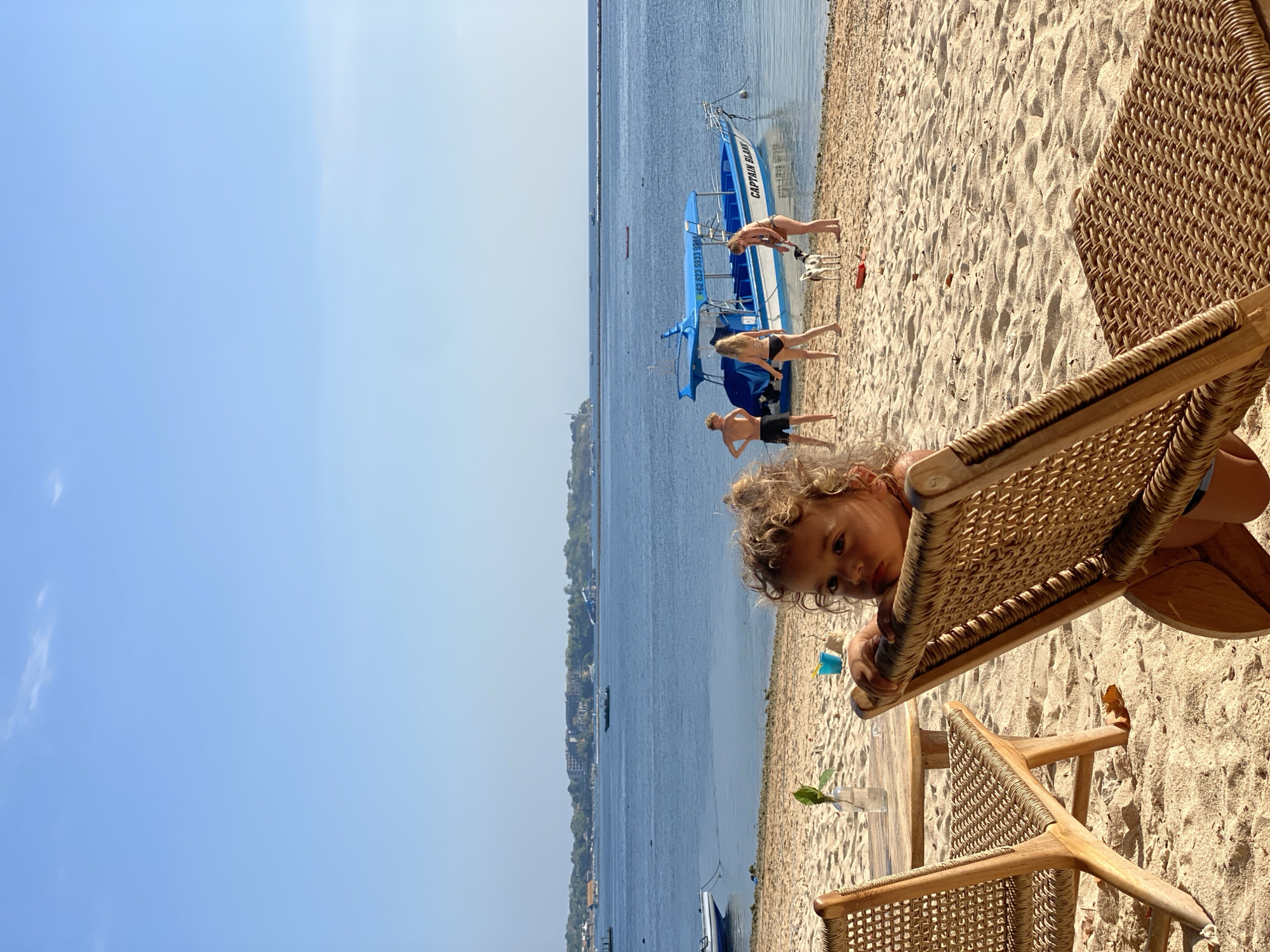 The image depicts a beach scene in Nusa Penida, Indonesia, on a sunny day with a clear blue sky. In the foreground, two woven lounge chairs and a small wooden table with a bottle of water and a plant are visible on the sand. A young child with curly hair is looking over the back of one of the chairs.
In the middle ground, several people are on the beach near a blue and white boat named "CAPTAIN BLANK." Two individuals are standing on the sand, one appears to be a young woman in a bikini, and the other is a male. Another young woman is interacting with a small dog near the water's edge. A blue bucket and a red object are also on the sand.
The background features the calm, blue ocean with several boats in the distance. The shoreline is lined with vegetation and buildings, suggesting a coastal town or resort area. The water extends to the horizon, meeting the clear sky.