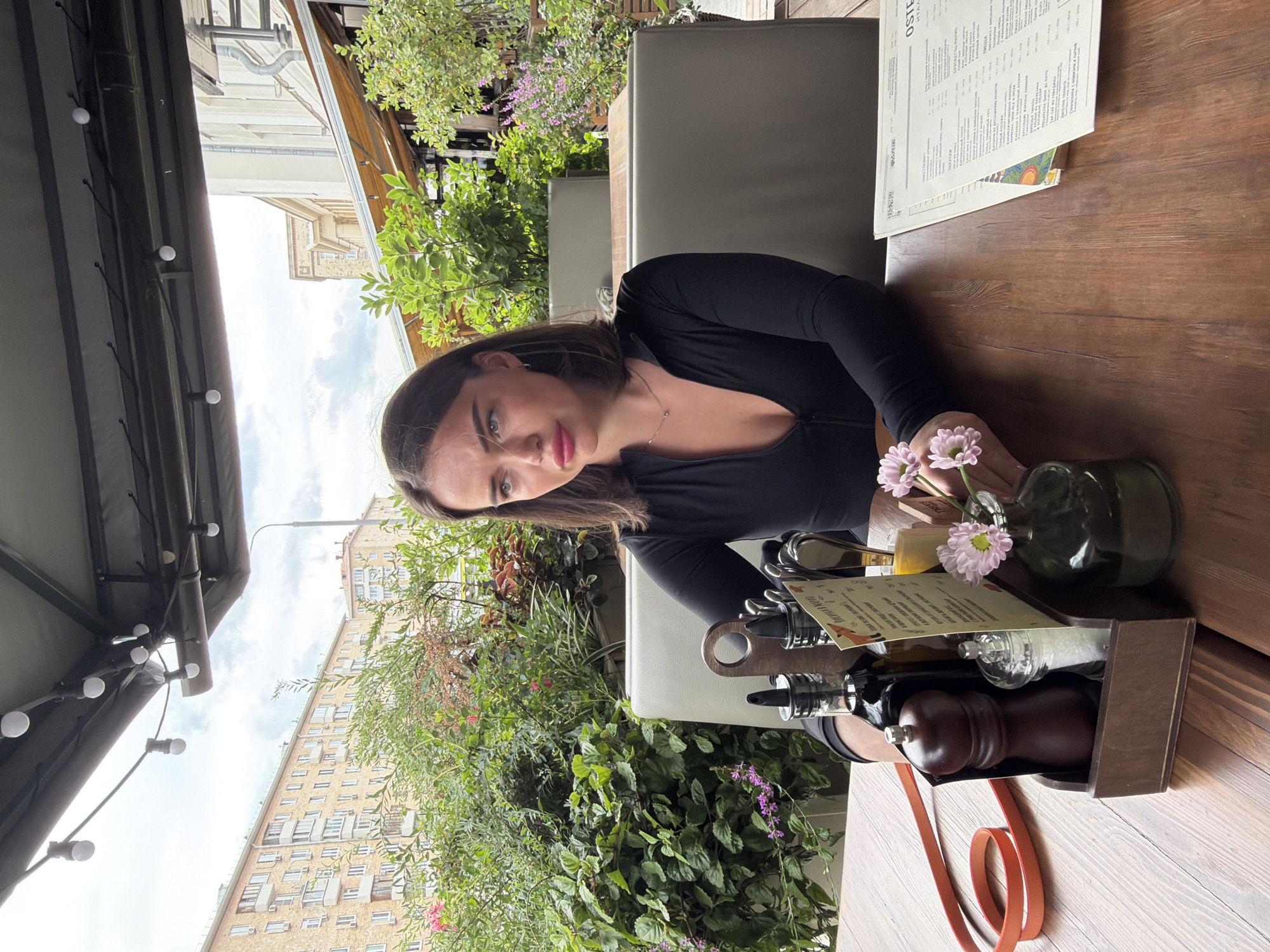 A young woman with dark hair and blue eyes is seated at a wooden table on an outdoor patio, likely a restaurant. She is wearing a black, form-fitting long-sleeved top with a deep V-neck. She is looking off to her right with a slightly pensive expression. On the table in front of her is a wooden caddy containing a pepper grinder, a salt shaker, and two bottles, one appearing to be olive oil. A menu is partially visible to her right, along with a small vase holding two light pink flowers. Behind the woman, a lush green plant wall separates the patio from the street, where multi-story residential buildings and an older building with awnings are visible. The sky is overcast.
