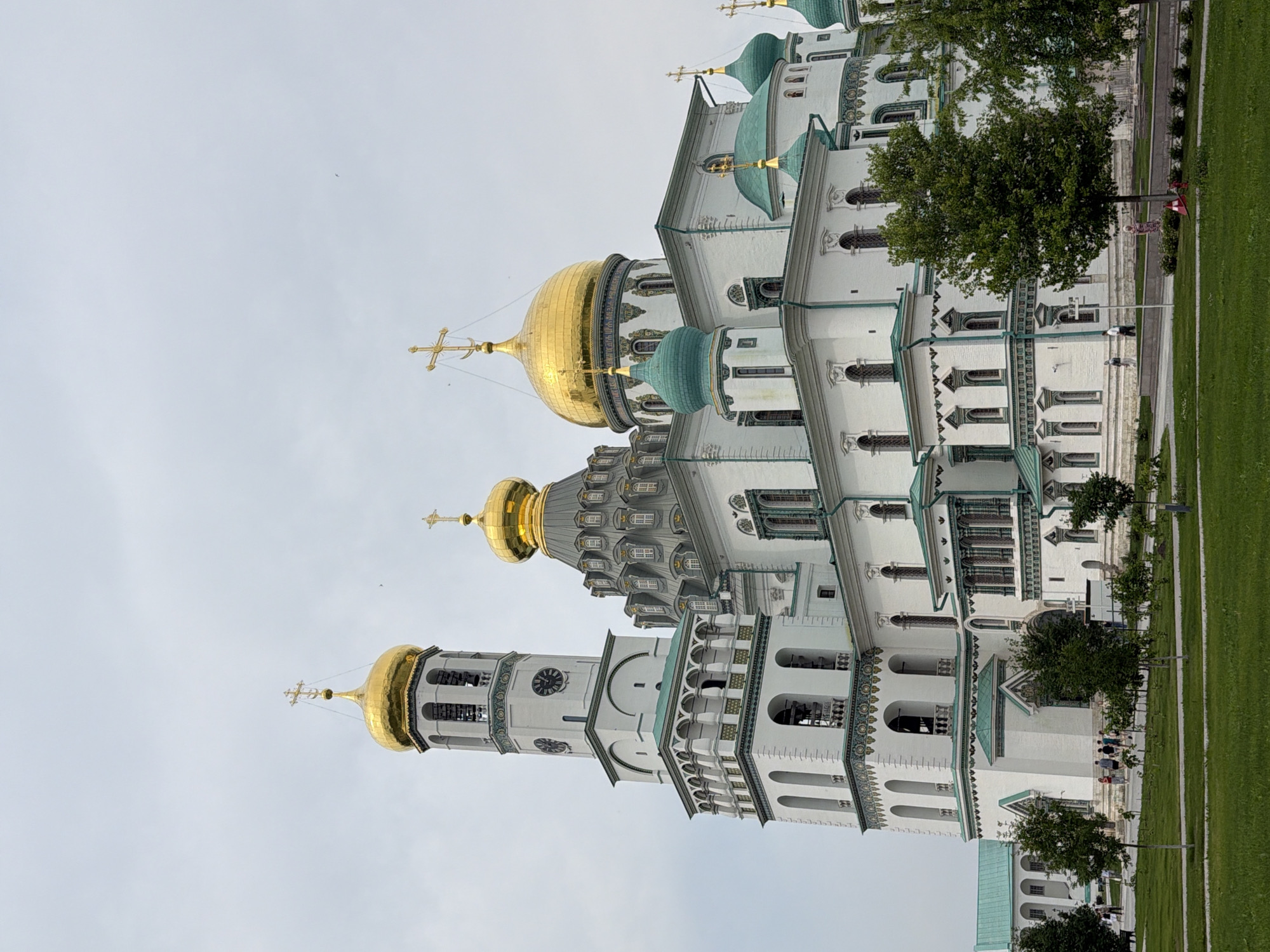The image captures the New Jerusalem Monastery, a prominent religious complex located in Istra, Russia. The main structure is a large, white building with multiple golden domes topped with crosses, and several smaller green domes. A tall bell tower with a clock face is visible to the left of the main structure. The building exhibits intricate architectural details with decorative trim along the rooflines and window frames. In the foreground, a well-maintained green lawn extends outwards, with several trees of varying sizes scattered across it. A paved pathway runs along the edge of the lawn. In the background, a few people can be seen walking on the pathway and near the monastery entrance. The sky is overcast and cloudy, creating a soft, diffused light.