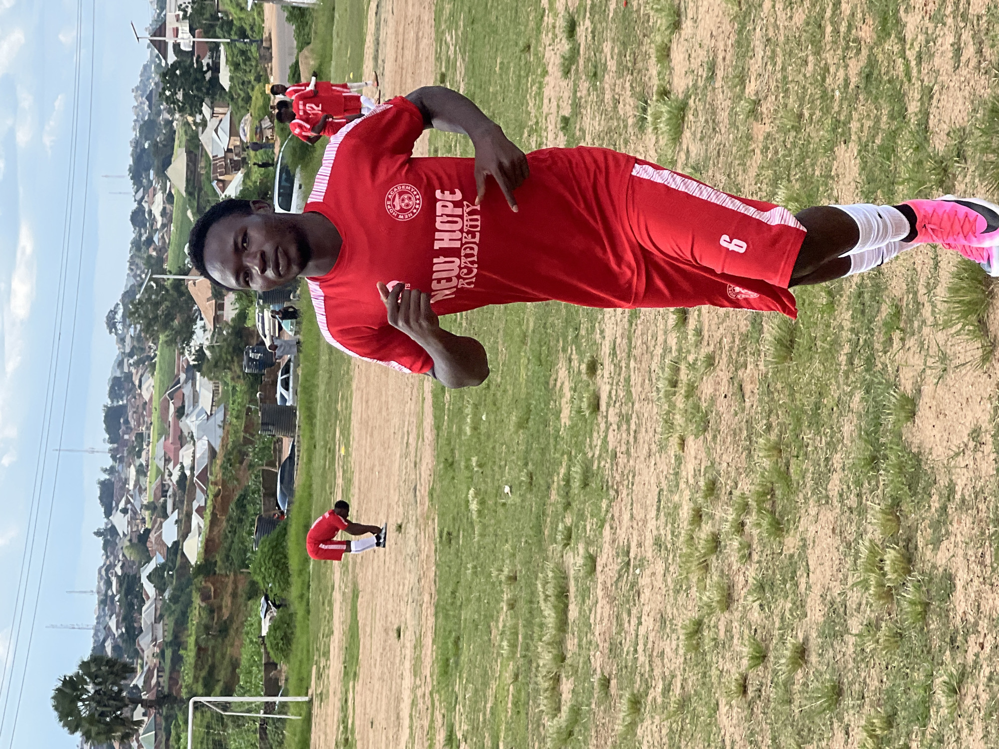In Jos, Nigeria, on a dusty football field dotted with sparse green grass, a young man in a red "New Hope Academy" jersey and shorts runs towards the camera. He wears pink soccer cleats and white socks. Another player in a red jersey with the number 12 is visible in the background, along with other teammates. The field overlooks a dense residential area with red-tiled roofs and scattered trees. A soccer goalpost is visible on the left side of the frame. The sky is overcast with a few wispy clouds.