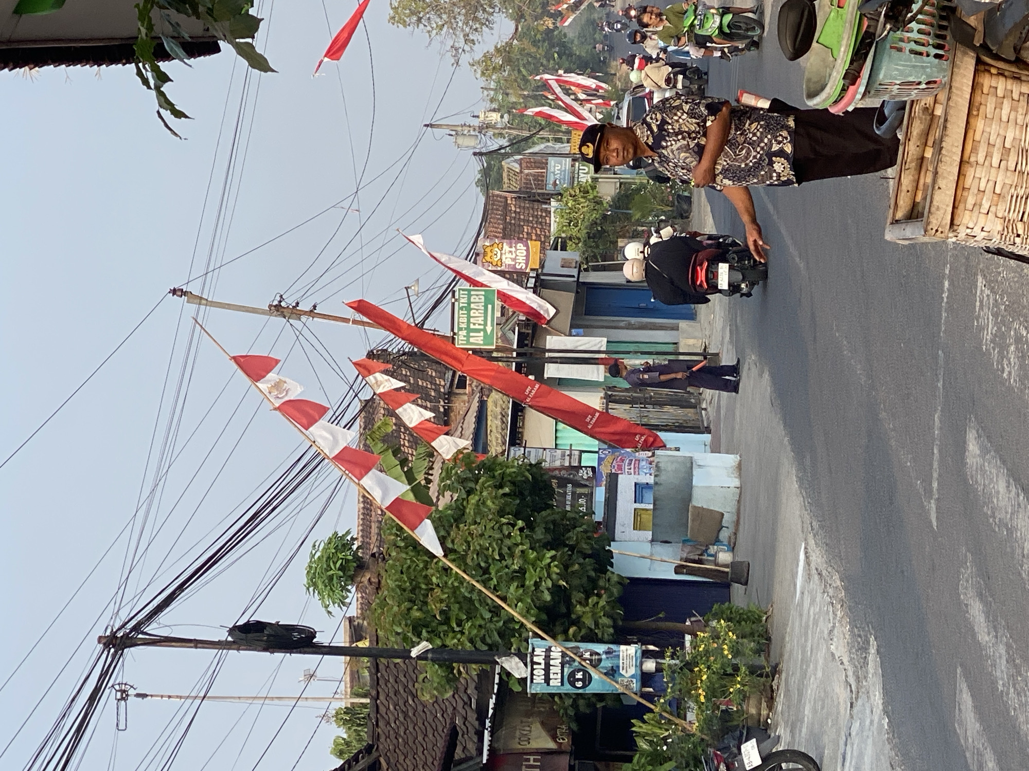 The image depicts a street scene in the city of Kasihan, Indonesia, adorned with red and white flags, likely for a national celebration. A man in a patterned shirt and dark pants stands on the right side of the street, looking towards the viewer. He is wearing a dark cap. Behind him, traffic, primarily motorcycles, moves along the road. Several buildings line the street, featuring tiled roofs. One building displays a sign for "TPA-KBIT-TKIT AL FARABI". The street is paved, with white pedestrian crossings visible. Power lines crisscross above the street and buildings. On the left side of the image, a tree with green foliage is prominent. A motorcycle is parked on the left side of the road, with part of its wheel and license plate visible. In the foreground, a wicker basket sits on what appears to be part of a motorcycle. The sky is clear and light blue.