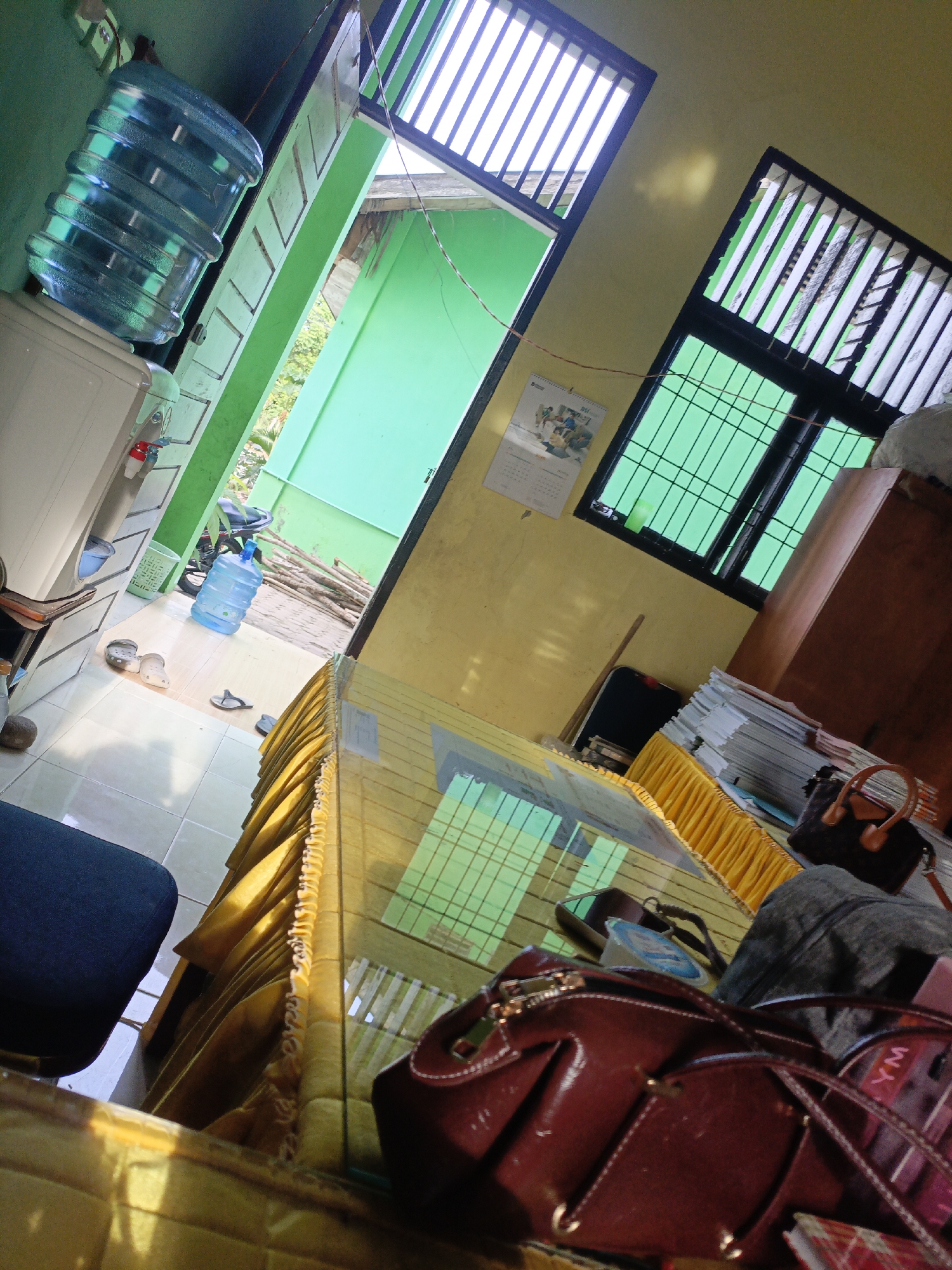 The image depicts an interior scene, likely a room, with a tilted perspective. A large blue water jug sits atop a white water cooler unit. To the left, a partially open green door reveals an outdoor area with green walls and what appears to be a pile of wooden poles. Through another doorway, a window with green-tinted glass and black bars is visible. A calendar hangs on the yellow wall above the window.
In the foreground, a glass-topped table is covered with a yellow tablecloth. Several items are scattered on and around the table, including a dark blue chair on the left, a dark brown handbag with tan straps, a greyish bag, and a red handbag in the lower right corner, featuring gold-colored hardware and white stitching. A few pairs of flip-flops and clogs are on the floor to the left of the table.
The environment suggests a practical, possibly domestic or small office setting in Lhokseumawe, Indonesia, characterized by the functional water cooler and the presence of personal belongings. The lighting suggests it is daytime, with sunlight casting shadows. The overall impression is one of an occupied space with items left out, indicative of ongoing activity or recent use.