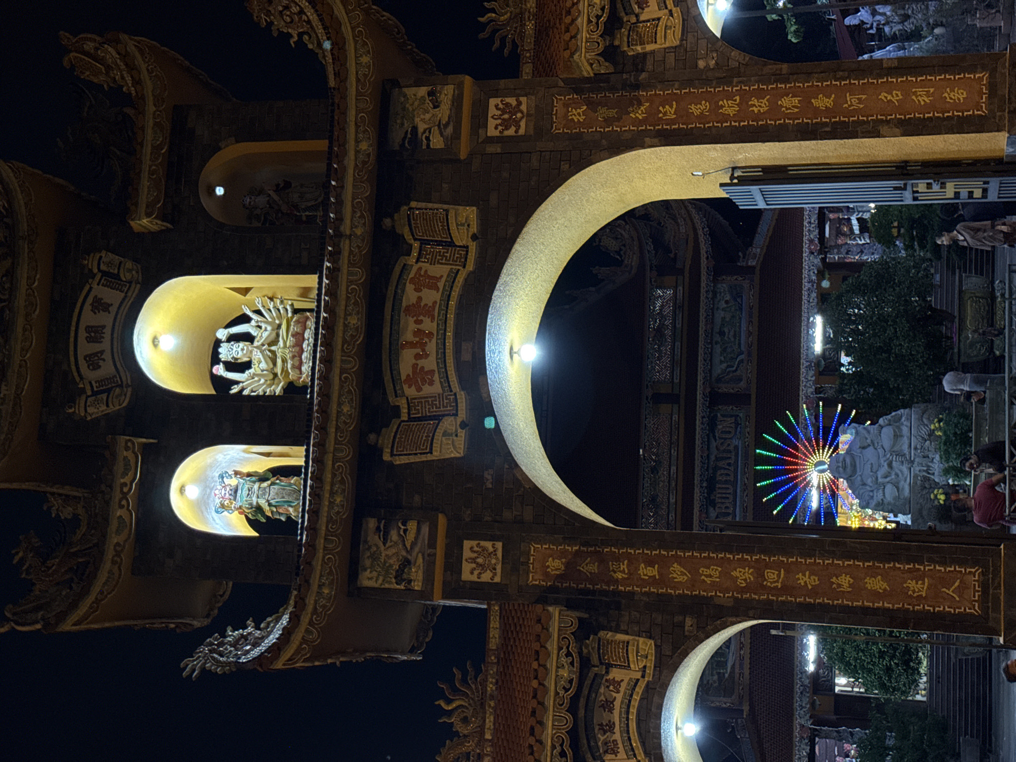 The image shows a night view of a temple entrance in Đà Nẵng, Vietnam. The temple gate is ornate with carvings, gilded details, and inscriptions in Chinese characters. Niches are illuminated to display statues of deities. The architecture features archways. Behind the entrance, a statue is illuminated with a colorful, radial light display. There are people visible in the background.