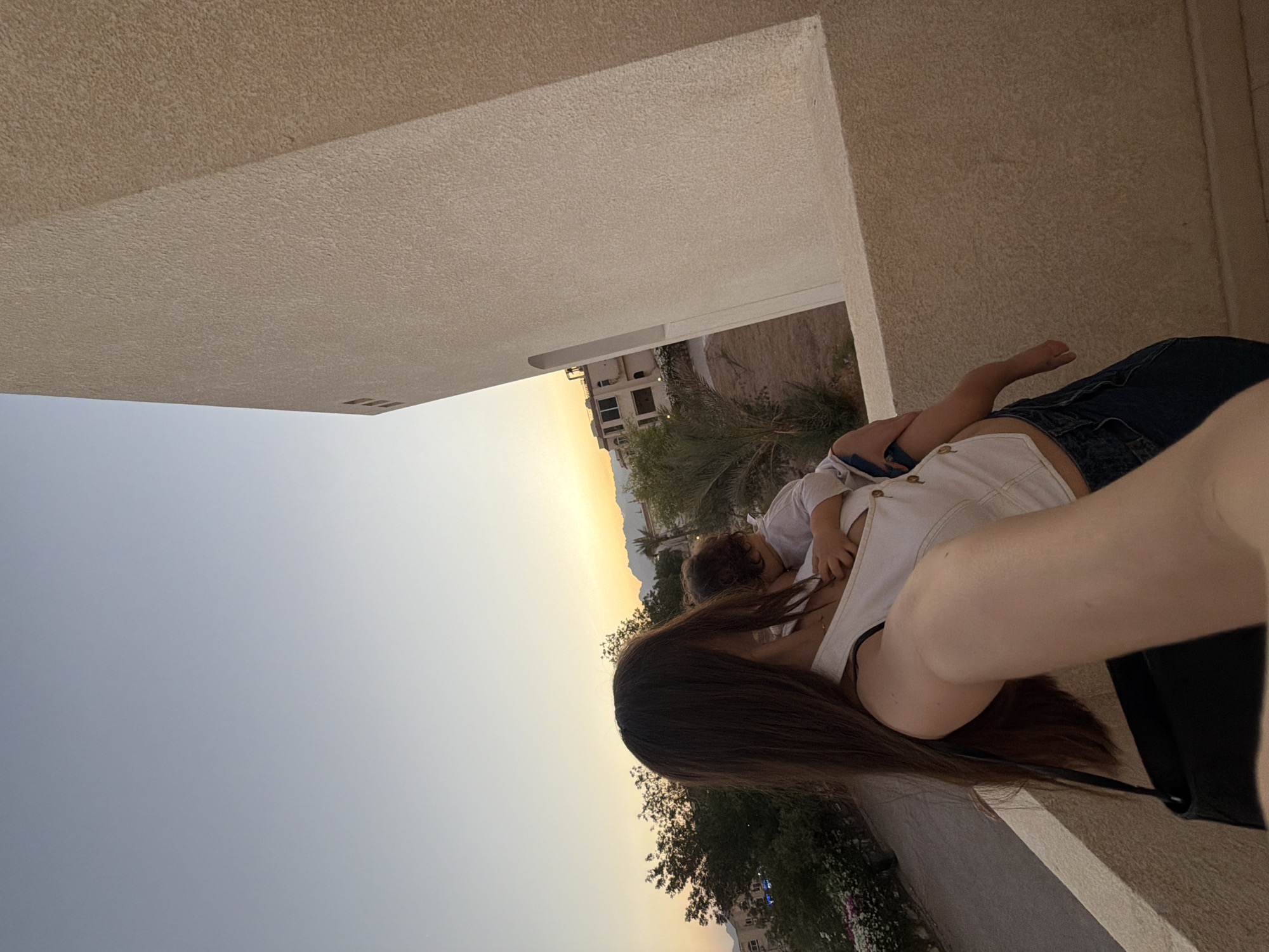 A woman with long brown hair is holding a child. The woman is wearing a white sleeveless top and blue jeans. The child is wearing a white outfit and is nestled in the woman's arms. They are standing on a balcony with a beige textured wall on the right side of the frame. In the background, there are buildings, trees, and mountains. The sky is a gradient of pale blue to orange, indicating either sunrise or sunset. The scene appears to be in a city, possibly Sharm El Sheikh, Egypt, given the architecture and natural landscape.