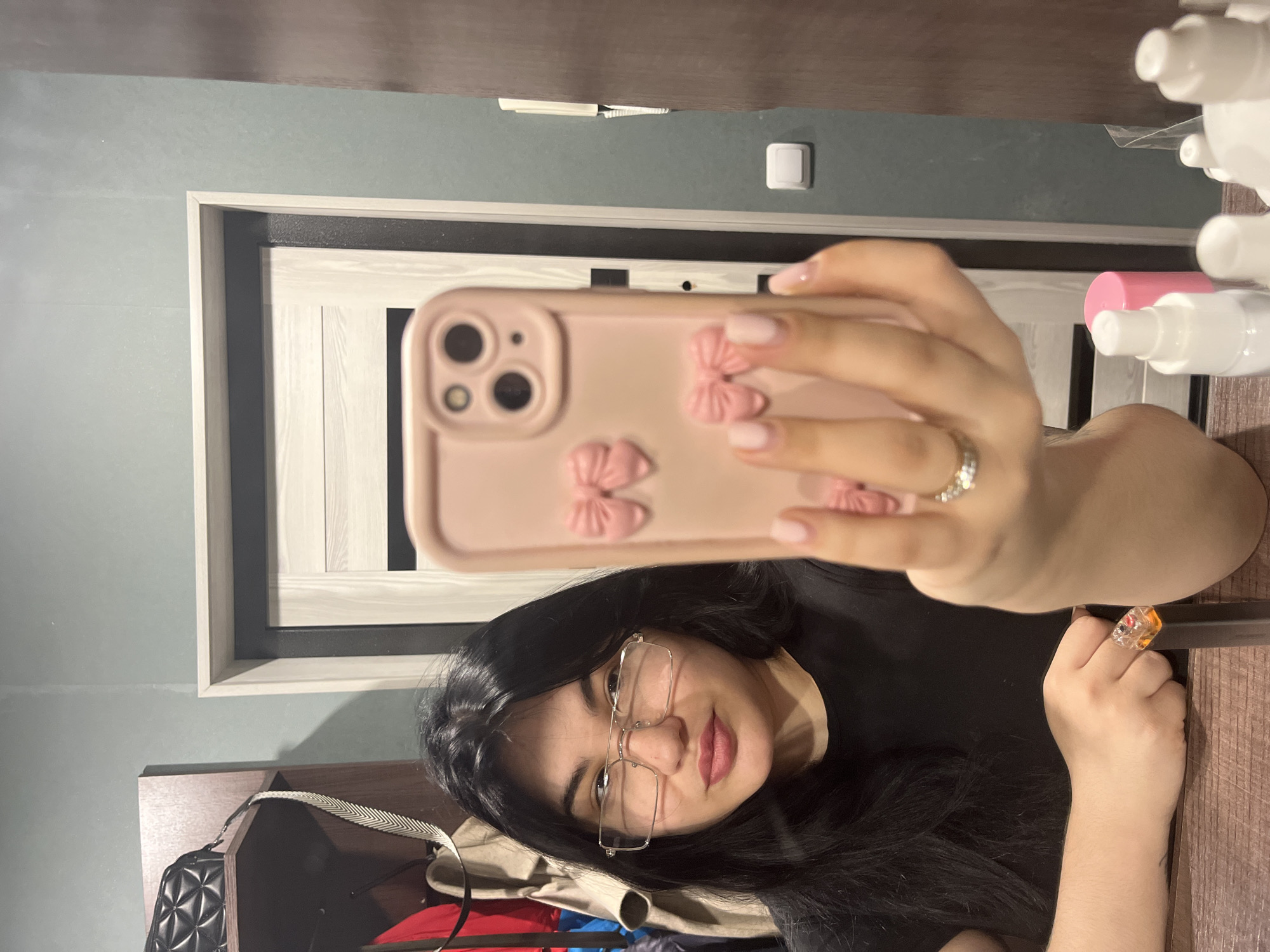 The scene depicts an indoor residential environment, consistent with a dwelling in Chaikovsky, Russia. A single female individual is visible, captured in a mirror reflection. She has long, dark hair and wears rectangular-framed eyeglasses and a black short-sleeved top. Her fingernails are painted light pink, and a ring with multiple small stones is on her left ring finger. The female is actively taking a mirror selfie, holding a light pink smartphone with a decorative case featuring three prominent pink bows in her right hand. The phone's multi-lens camera array is visible. Her left arm rests on a wooden surface in the foreground.
The reflected background shows a room with walls painted a dark teal or green-blue color. A light wood-paneled door with dark interior trim and a light outer frame is centered behind the subject's reflection. To the left, a wooden shelving unit contains various hanging garments, including red and beige items, and a black quilted shoulder bag. To the right of the door, a white light switch and a white intercom unit are mounted on the wall. The wooden surface in the foreground, presumably a dresser or table, holds several small white spray bottles and one bottle with a pink cap. An orange and clear electronic device with visible internal components rests on this wooden surface to the subject's left.