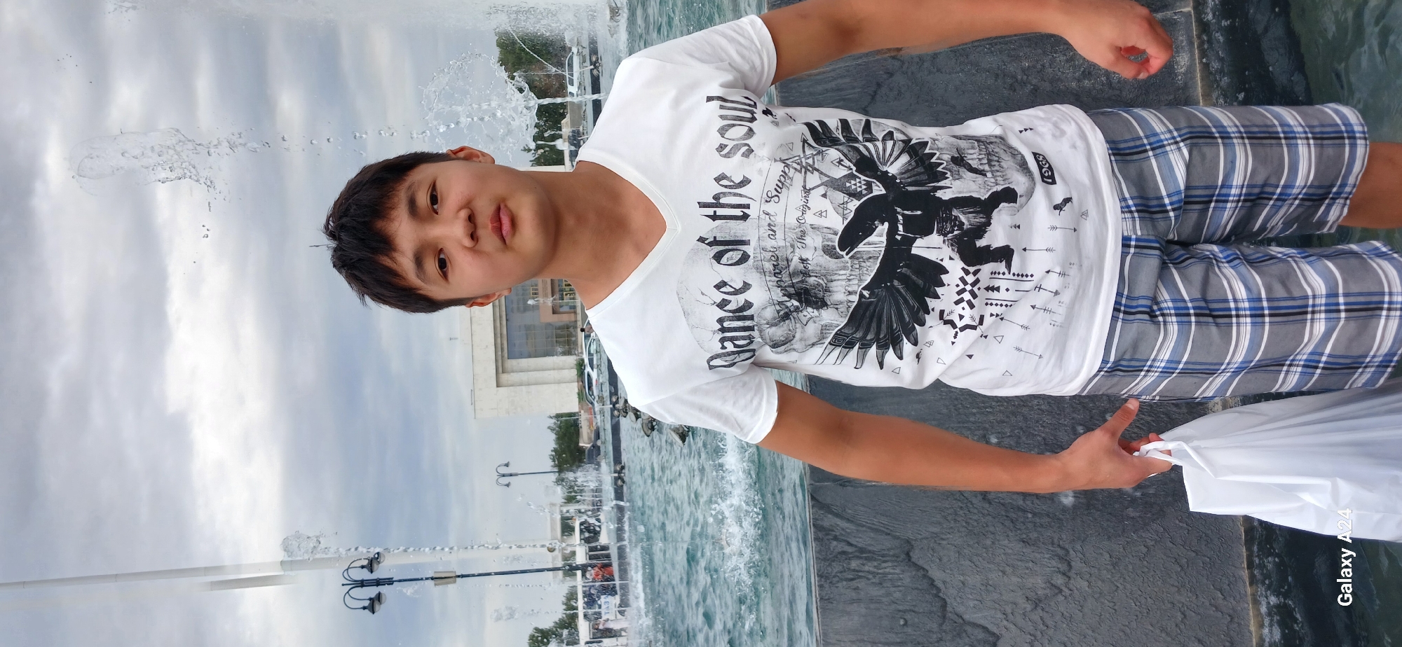 A young man with short black hair and brown eyes stands in front of a fountain in Bishkek, Kyrgyzstan. He is wearing a white V-neck t-shirt with a graphic print and plaid shorts. He is holding a white plastic bag in his left hand. The fountain is spraying water, and there are buildings and cars in the background. The sky is cloudy. The image appears to be taken during the daytime.