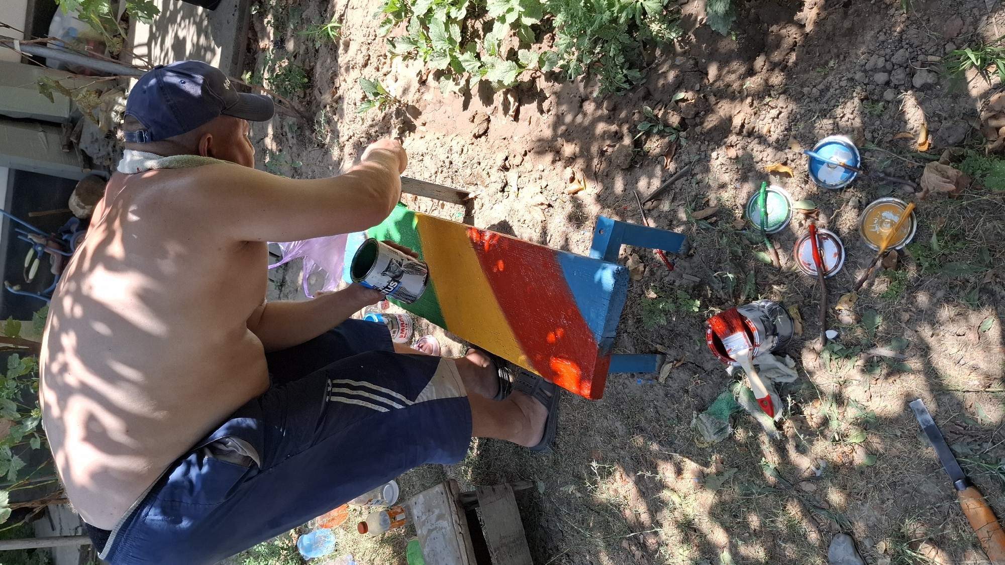 In Шұбарсу, Kazakhstan, a shirtless individual with a blue baseball cap and light-colored towel around their neck is seen painting a wooden bench outdoors. The bench is sectioned into four distinct color blocks: blue at the bottom, red, yellow, and green at the top. The person is holding a can of green paint and appears to be in the process of applying it to the bench. Several open paint cans in various colors (red, yellow, blue, and green) are scattered on the ground around the bench, along with paintbrushes and a chisel. The environment is a dirt yard with dry grass, vegetation, and a building or structure in the background. Sunlight casts shadows from overhead foliage onto the scene.