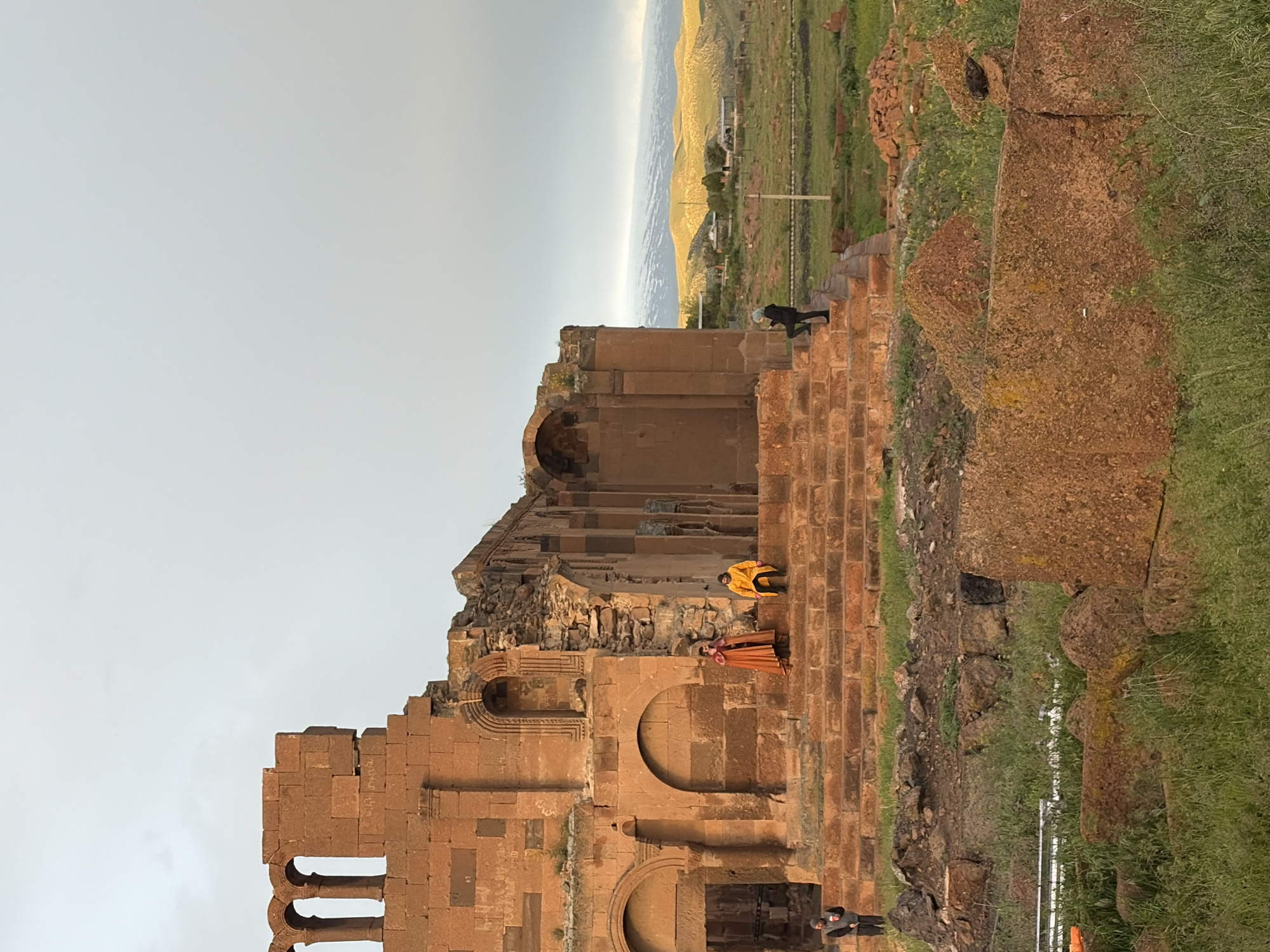 The image depicts the ruins of Ani Cathedral in Anipemza, Armenia. The structure is predominantly brown stone with arched openings. Three people are visible: one dressed in orange on the steps, one in yellow above them, and one in black on the top of the wall, all situated within the ruins. A landscape with hills and sky are visible in the background. Grass and some debris are present on the ground surrounding the structure.