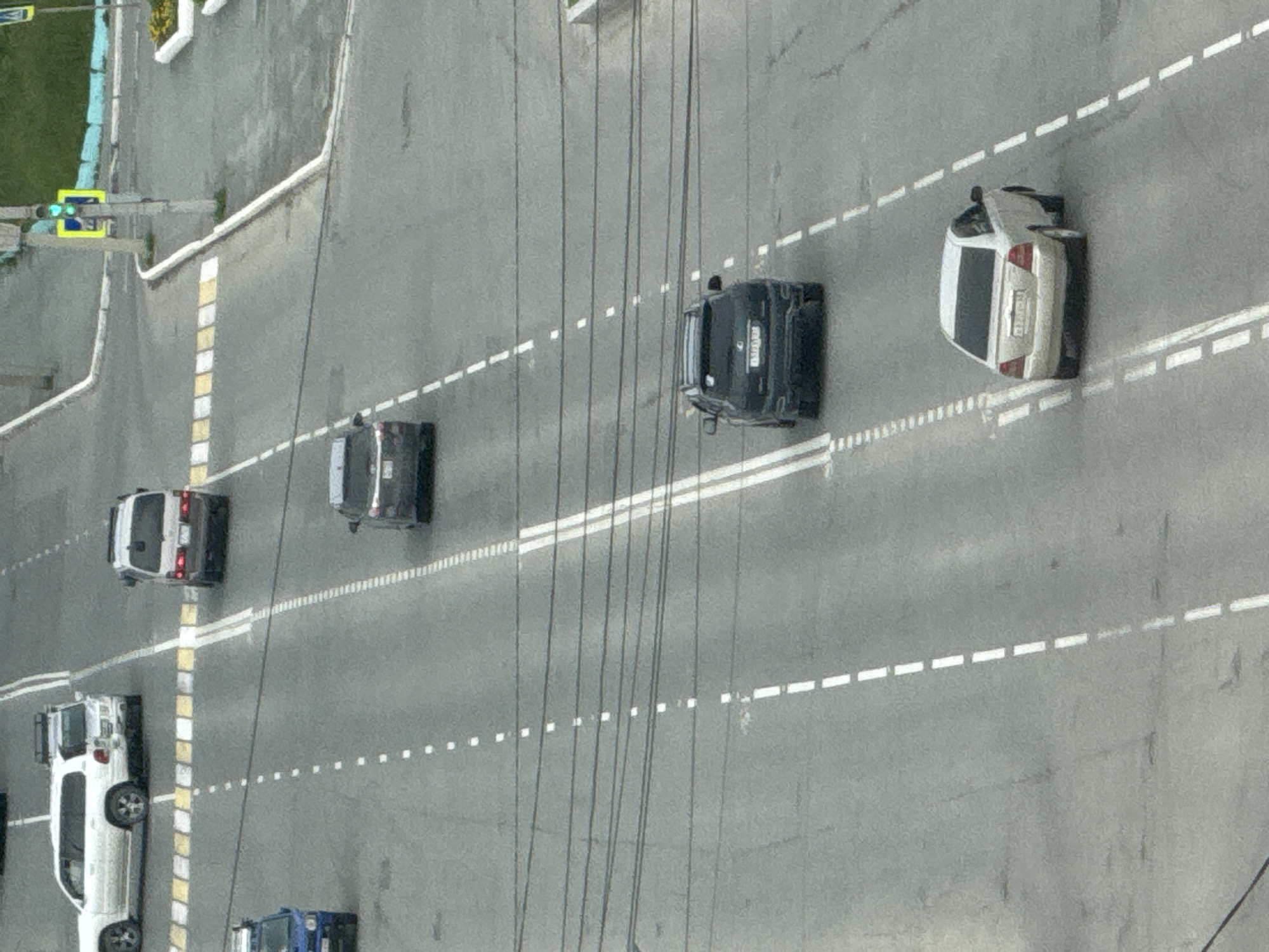 The image shows a multi-lane asphalt road with various vehicles. In the foreground, a white sedan is moving away from the viewer, traveling in the right lane. Behind it, a dark colored SUV is also in the right lane, following the sedan. Further up the road, a dark colored sedan is in the left lane. Even further up, a dark colored van is in the right lane. In the top left of the image, a white SUV is traveling in the left lane, and a blue vehicle, possibly a van, is in the left lane. The road is marked with white dashed and solid lines, indicating lanes and crosswalks. Overhead power lines are visible stretching across the scene. In the upper right quadrant, a traffic light is visible, showing a green signal, and a yellow pedestrian crossing sign is present. The environment appears to be an urban setting, consistent with a street in Kholmsk, Russia, given the typical architecture and road infrastructure. The weather appears to be overcast or hazy, contributing to the muted colors of the scene.