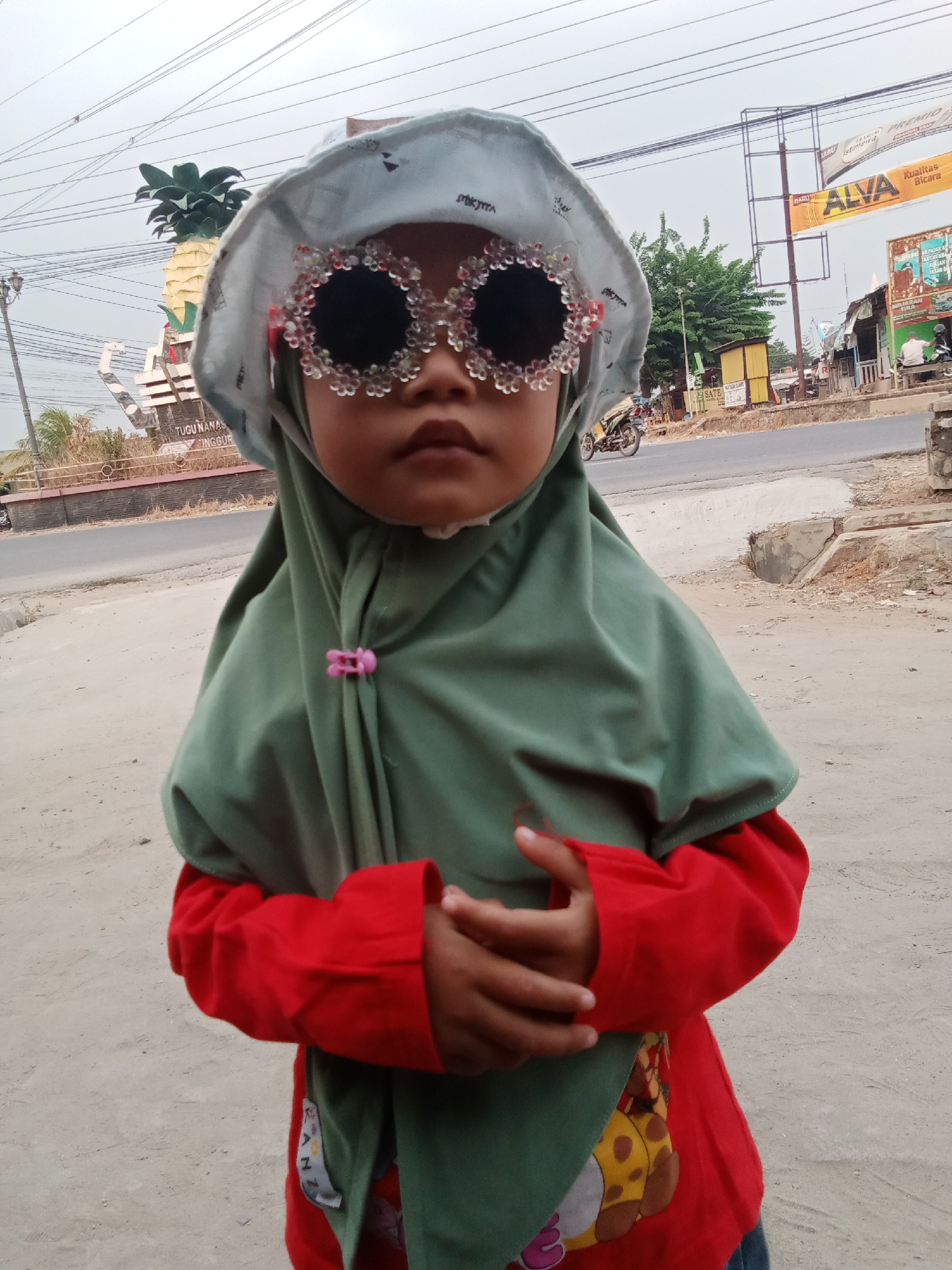A child wearing a light blue bucket hat adorned with sparkly pink and white embellishments and oversized, circular, jeweled sunglasses stands in front of a street scene in Toto Katon, Indonesia. The child is also wearing a green hijab, a red long-sleeved shirt, and a red shirt with a cartoon giraffe print. Their hands are clasped in front of their body. In the background, a large pineapple monument labeled "TUGU NANAS DINGGUR" is visible, along with various shops, a road with passing vehicles, and overhead power lines. The weather appears overcast.
