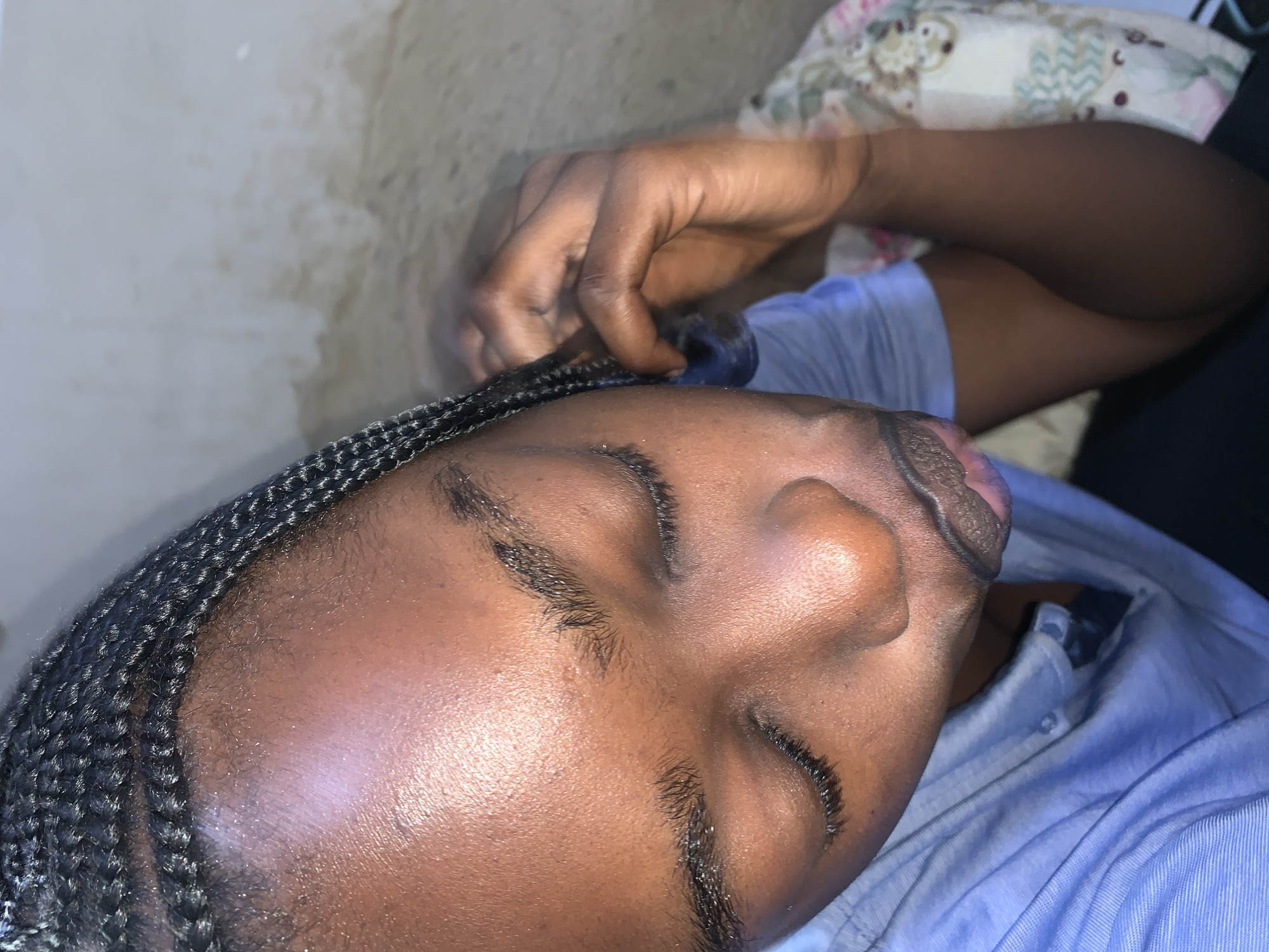 A close-up shot, taken from a high angle, shows a person's face and upper body. The individual has braided dark hair, and their eyes are closed. They are wearing a light blue collared shirt. Their right hand is raised and touching their hair. The background appears to be a wall with some discoloration. This image was captured in Rafin Par, Nigeria.