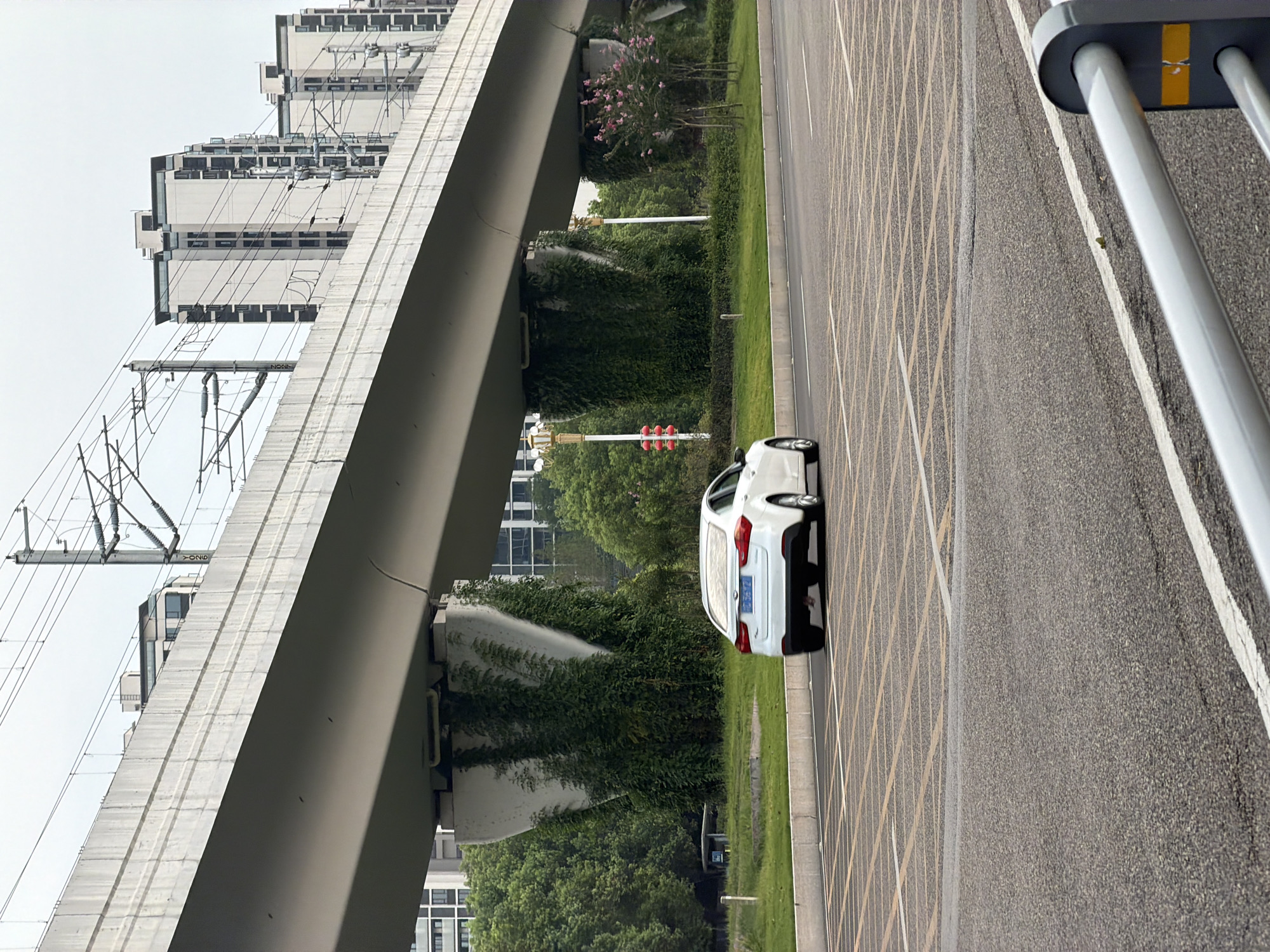 A white sedan is driving on a road in front of a raised railway track in Jiangjin, China. The railway track is supported by concrete pillars overgrown with green vines. In the background, modern apartment buildings are visible. The sky is overcast. The road has yellow and white markings indicating parking spaces or a special zone. A grey metal guardrail is in the foreground.