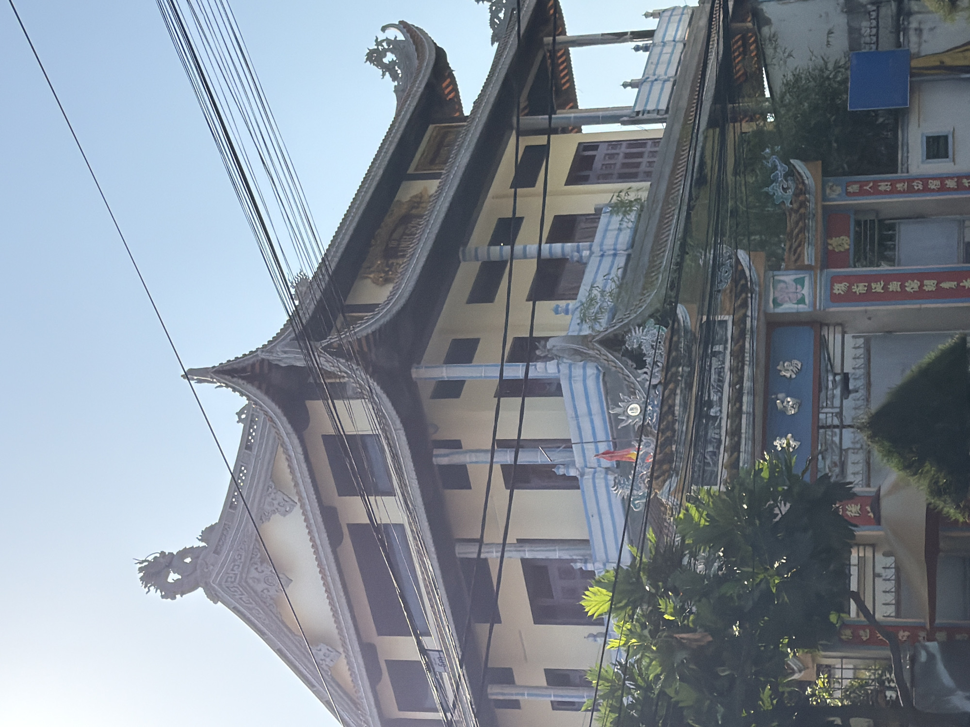 The image shows a multi-story building with traditional East Asian architectural elements, likely a temple or pagoda, situated in the city of Nha Trang, Vietnam. The building has a distinctive curved roof with ornate dragon carvings on the highest peak and along the eaves. The facade is painted in a light yellow color with dark trim around the windows and rooflines. There are multiple levels, each with recessed balconies supported by columns. The balconies have decorative blue and white striped railings.
In the foreground, lush green trees partially obscure the lower part of the building. The sky above is clear and blue, suggesting a sunny day. Electrical wires crisscross the frame, indicating the building is in an urban or suburban setting. Signs with Vietnamese characters are visible on a lower structure in front of the main building. The overall scene depicts a religious or cultural structure in its environmental context within Nha Trang.
