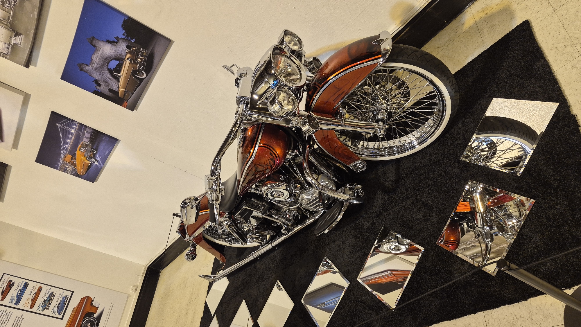 The image shows a customized orange and chrome motorcycle on display. The motorcycle is positioned on a black rug with a checkered pattern of white and black squares. Several square mirrors are placed on the rug, reflecting parts of the motorcycle. In the background, there is a white wall adorned with framed pictures of classic cars. One picture features a yellow car with the Golden Gate Bridge in the background, another shows a gold convertible in front of an archway. The scene appears to be indoors, possibly in a museum or exhibition space in Sacramento, California. There are no people visible in the image.