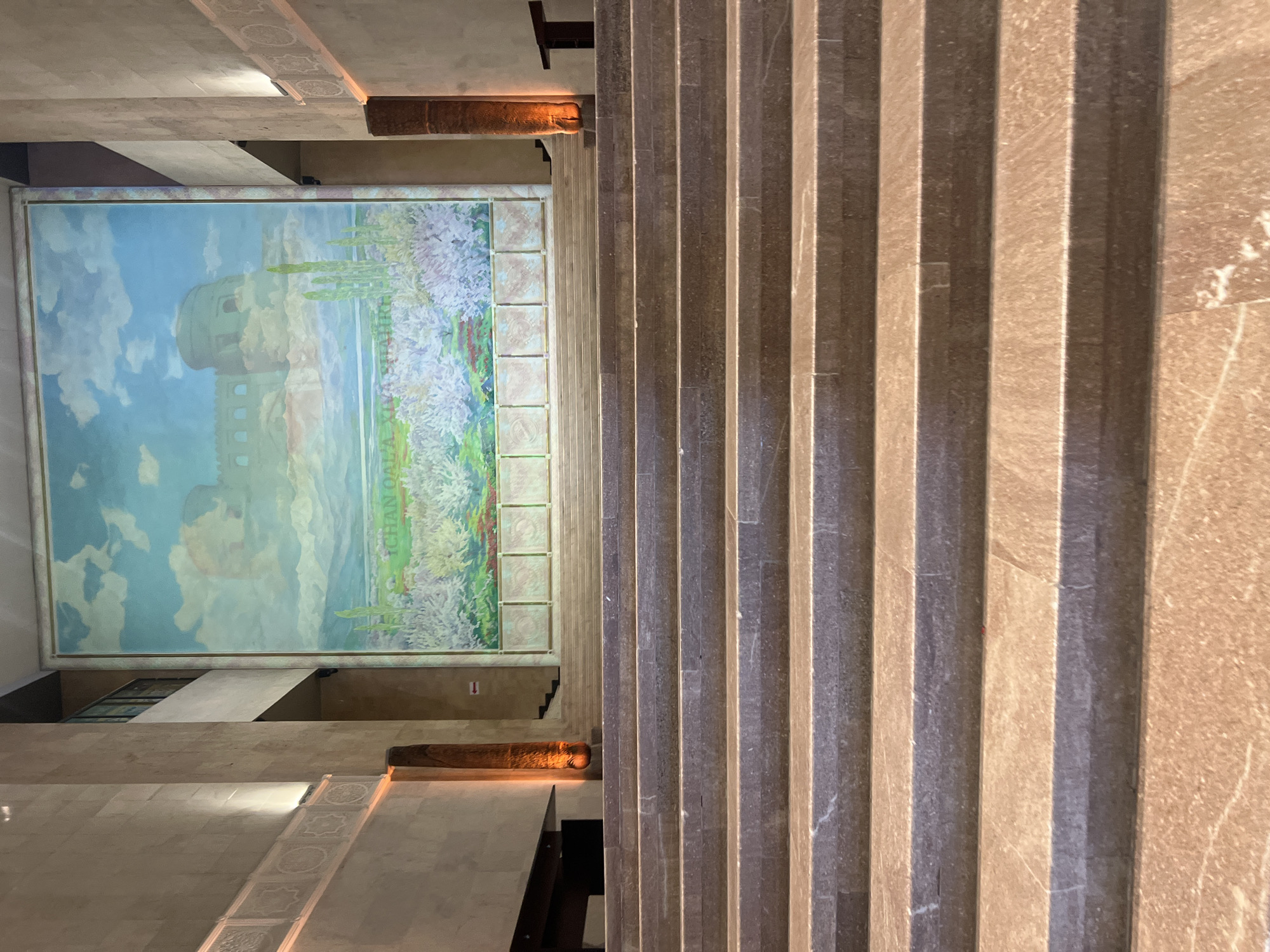 This is an interior, eye-level, medium shot of a grand staircase leading up to a large mural in the city of Unknown, Unknown. The staircase is made of polished, reddish-brown stone with subtle marbling. The steps are wide and evenly spaced. The mural on the wall above is a landscape painting depicting a historical fort or castle under a bright blue sky with white clouds, with a field of flowering trees or bushes in the foreground. The mural is framed by a decorative border. On either side of the mural are tall, rectangular pillars made of stone, with orange lighting emanating from their bases, creating an atmospheric glow. The walls flanking the staircase are also made of stone, with decorative friezes near the top. The overall impression is one of grandeur and historical significance, likely within a museum, government building, or similar public space. There are no people or animals visible in the image.