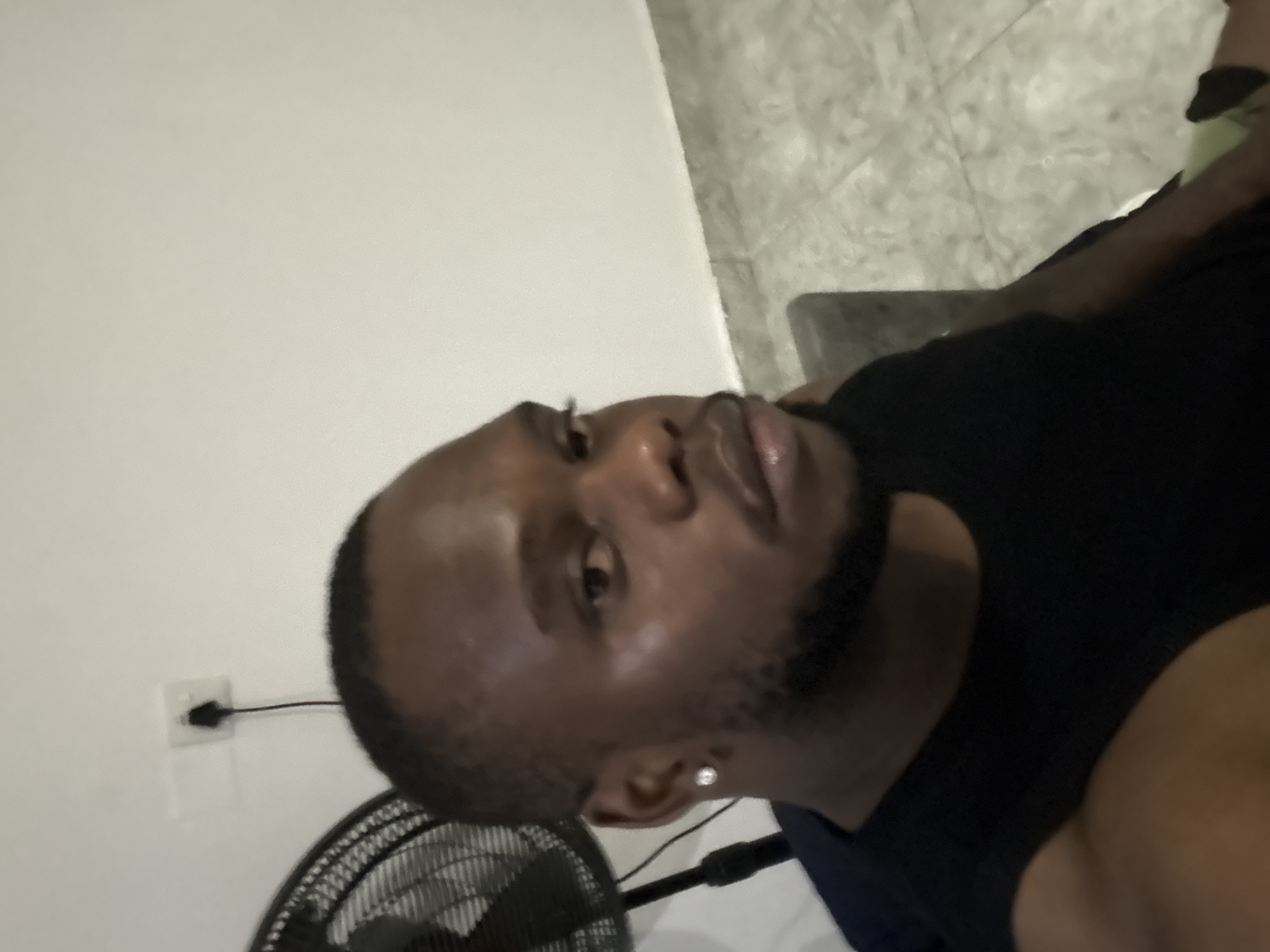 The image shows a man with dark skin, short dark hair, and a beard. He is wearing a black shirt and a small diamond earring. A fan with a black grill is in the lower left corner of the frame. A white wall is behind the fan, with a black electrical cord and a white outlet. The background includes a white wall and a tiled floor. The location is unspecified but is represented as the city of Iddo Tudunwada, Nigeria. The man is looking towards the camera.