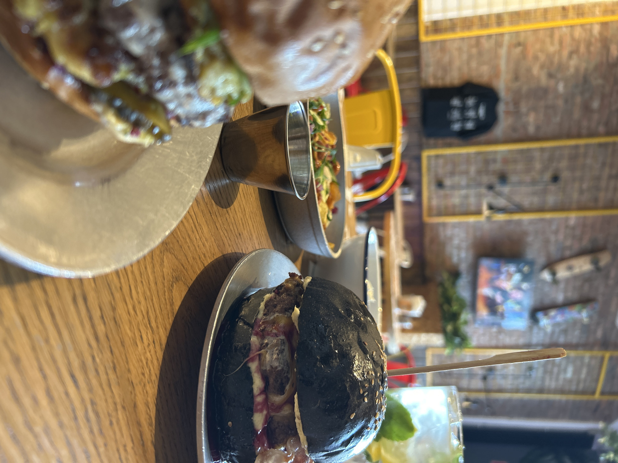 An eye-level, close-up shot shows two burgers and sides on a wooden table at a restaurant. In the foreground, a burger with a light-colored bun is mostly out of focus. To the right, a black burger with sesame seeds sits on a metal plate. It is pierced by a wooden skewer. Behind the burgers, a metal cup and a bowl of salad are visible. In the background, a brick wall with yellow metal accents and hanging skateboards and a t-shirt is out of focus. A drink with ice and a lime slice is also visible in the background.